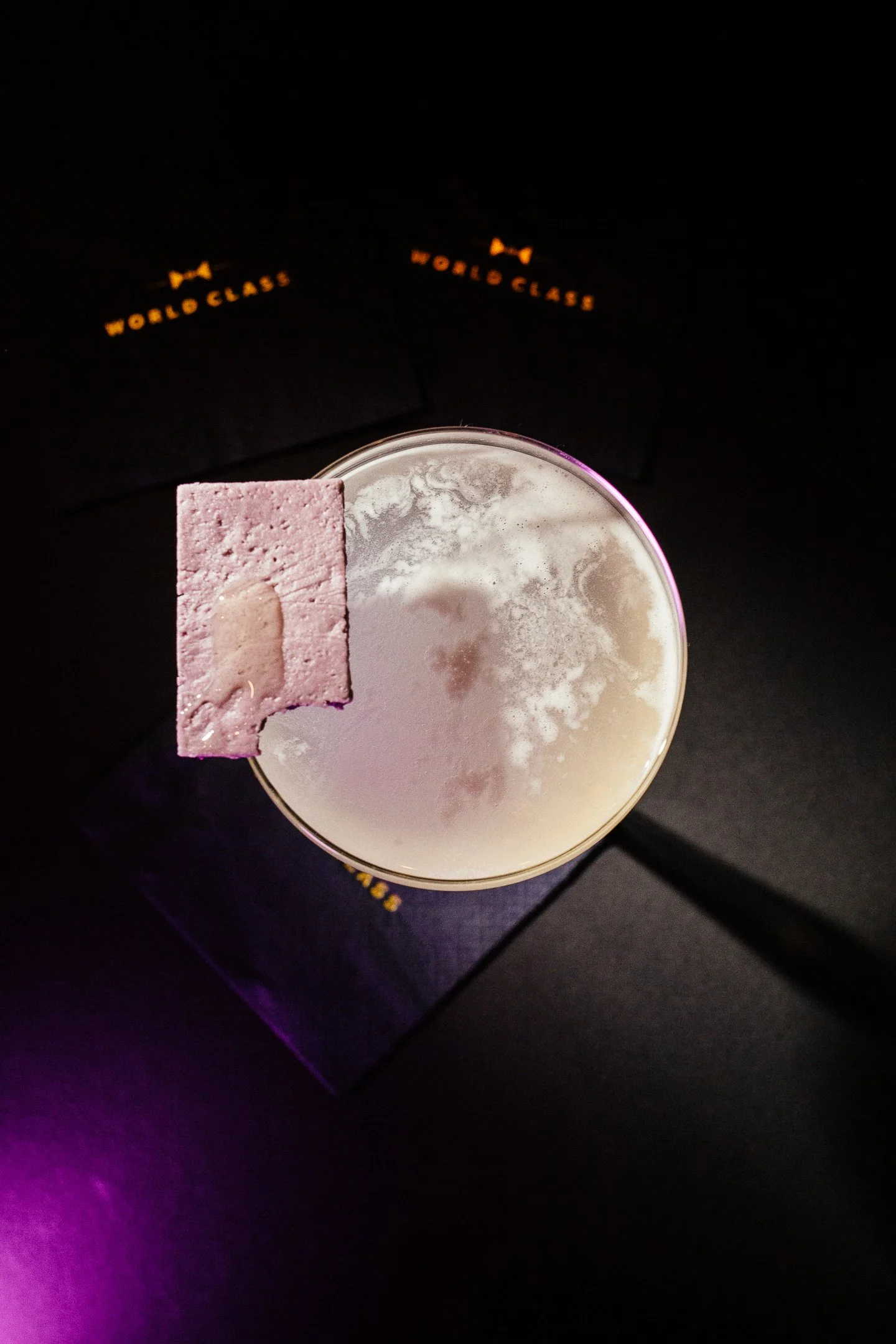 A glass of an alcoholic beverage with a pink wafer cookie on its rim, placed on a dark surface with a black box labeled 'World Class' in the background.