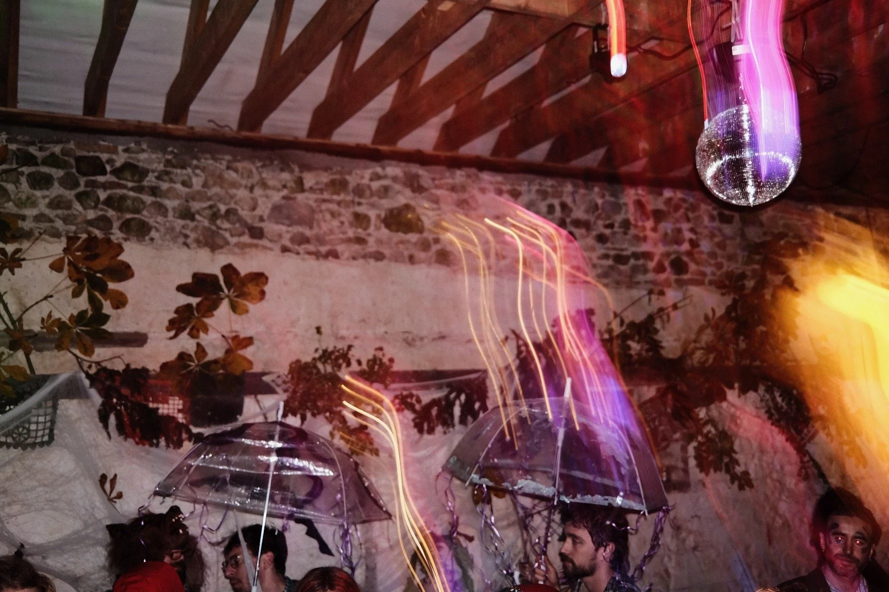 People at a party holding transparent balloons under a disco ball in a rustic venue with a wood ceiling and stone wall, colorful lights creating streaks.