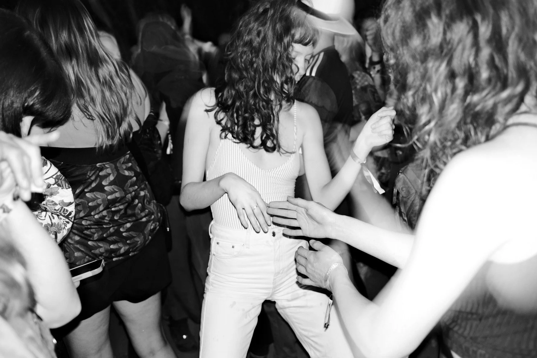 A woman with curly hair in a sleeveless top and white pants dancing at a party surrounded by multiple people.