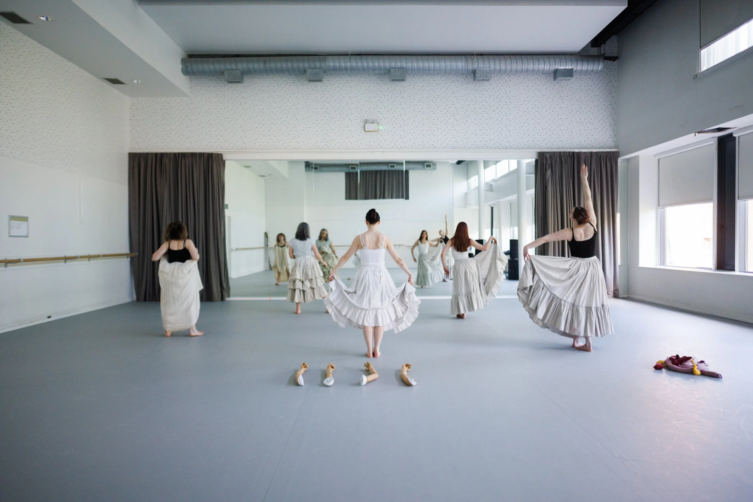 A group of women dressed in long, flowing skirts and tank tops practicing ballet in a dance studio with large mirrors and natural light from windows.