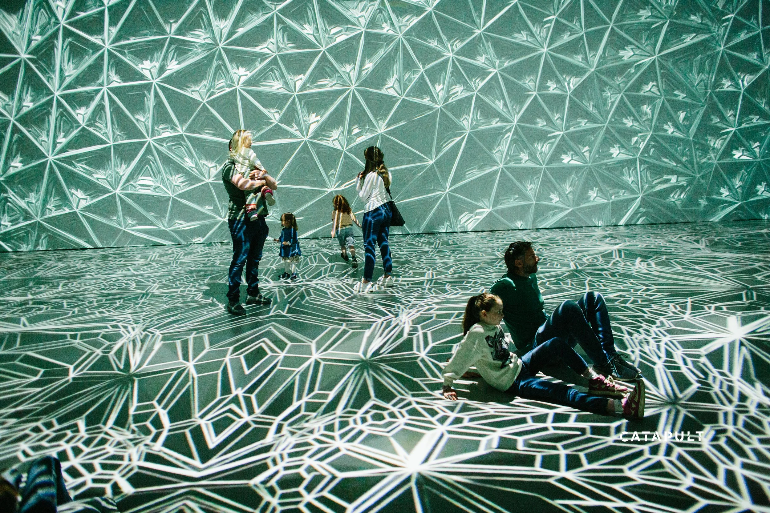 People sitting and standing inside a futuristic, geometric-patterned installation with illuminated walls.