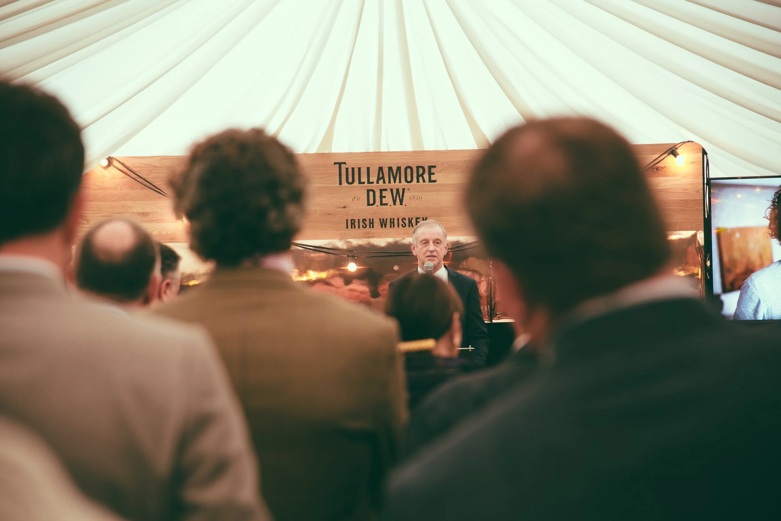 A speaker at an event standing in front of a Tullamore Dew Irish Whiskey sign, with an audience watching indoor presentation under a white, tent-like ceiling.