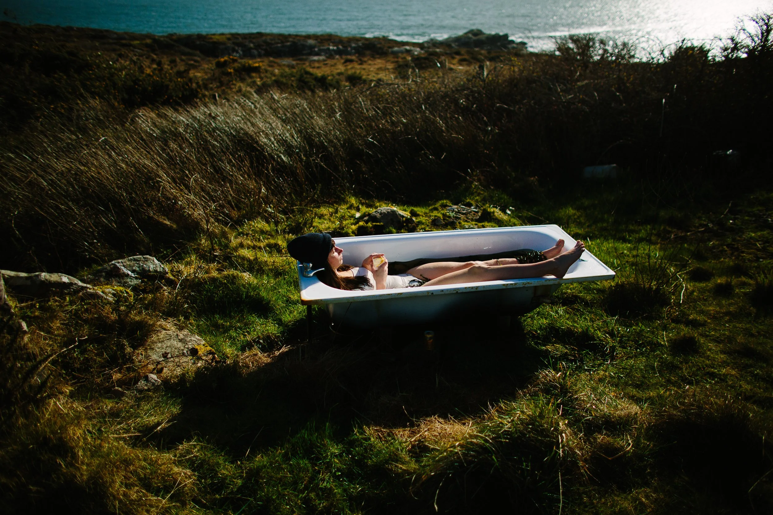 A woman lying in a bathtub outdoors on grassy terrain near a rocky hillside, holding a drink, with a body of water in the background.