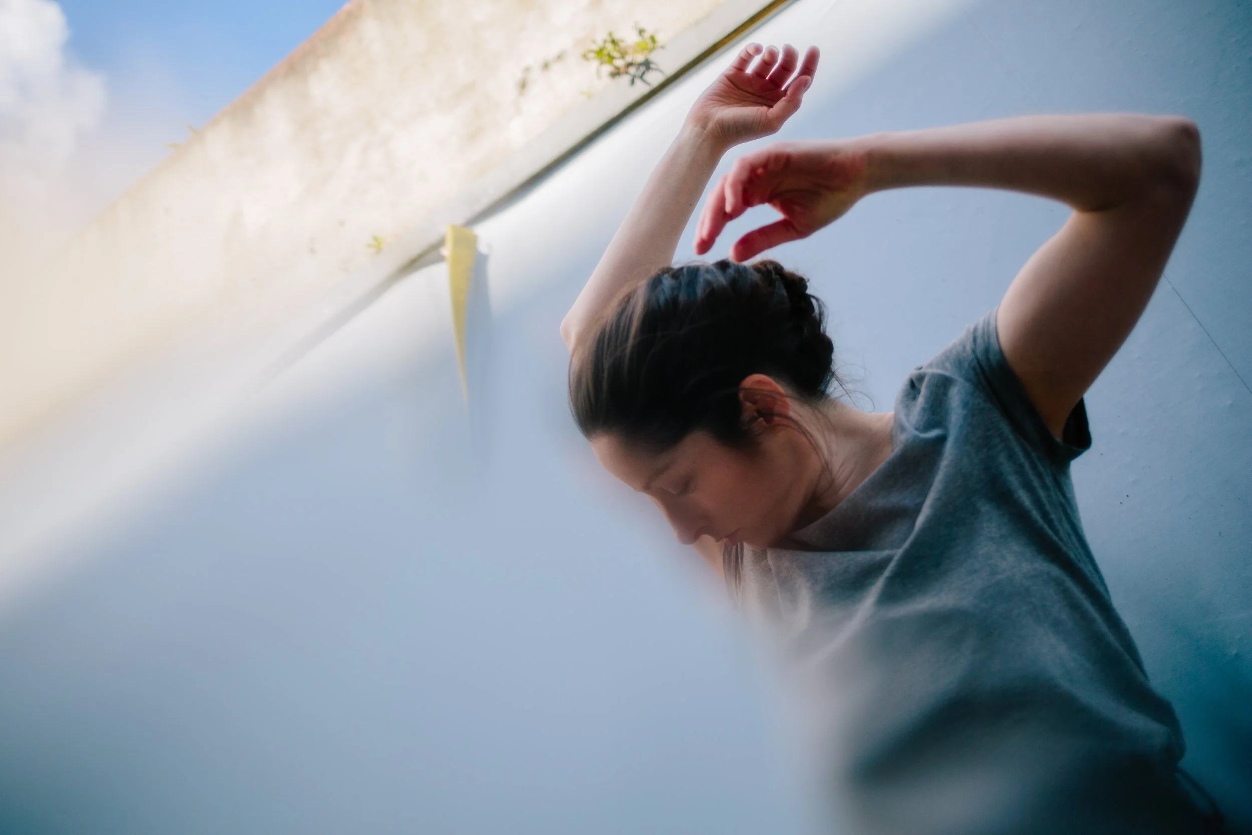 A woman practicing yoga or stretching indoors against a light blue wall, with her eyes closed and wearing a grey T-shirt.