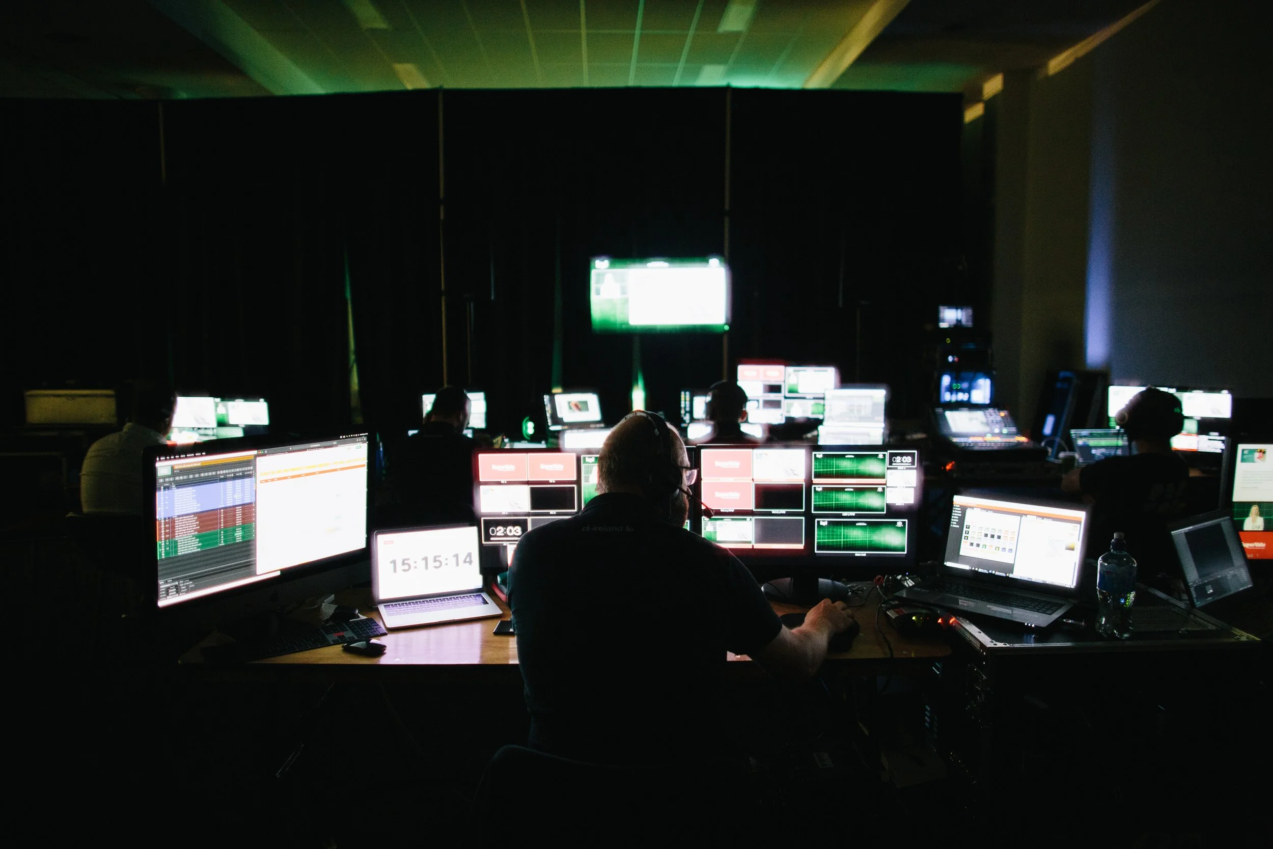 A darkened room with multiple people working at computer stations, illuminated screens, and large monitors displaying various data and graphics, suggesting a control room or broadcast studio environment.