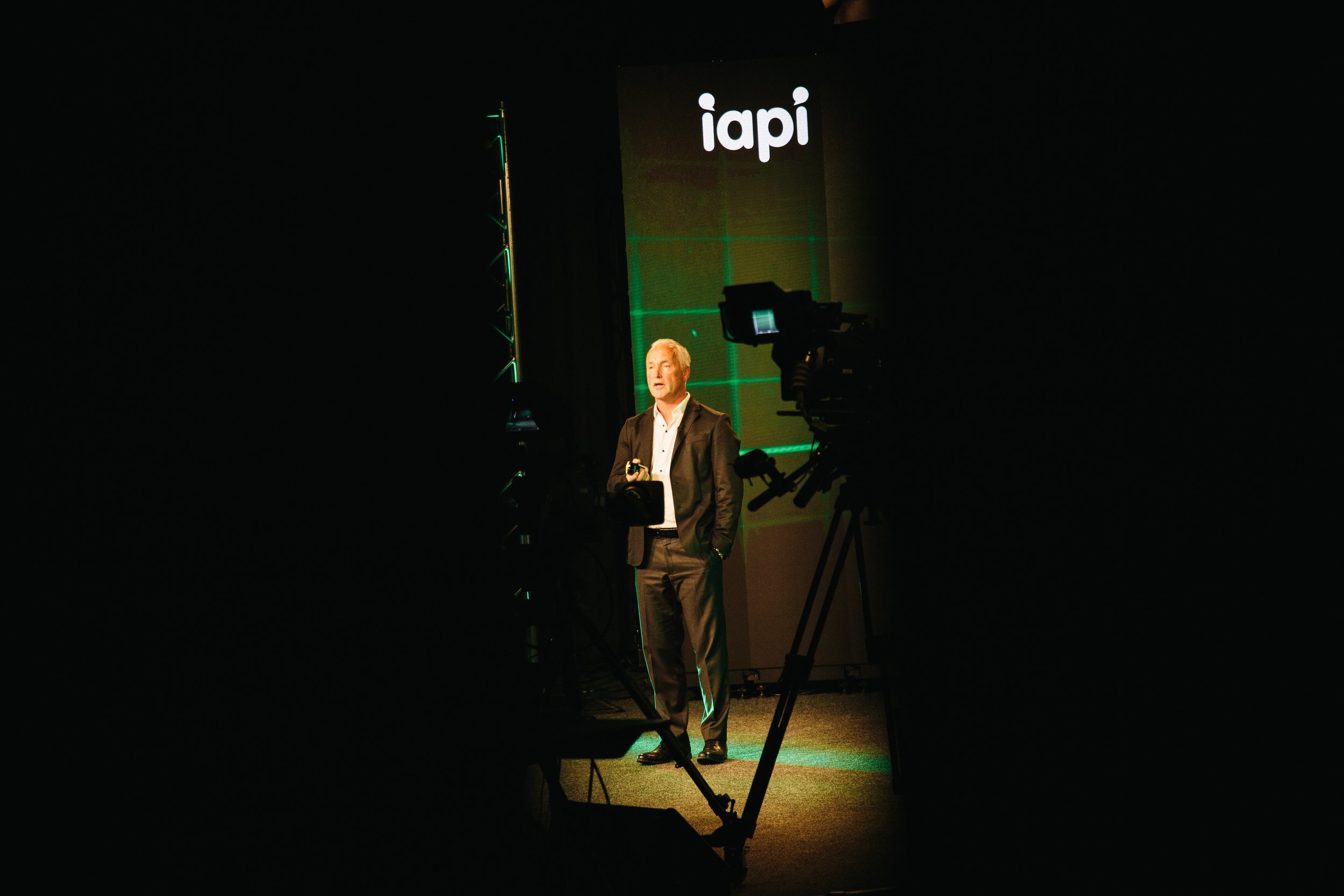 A man in a suit speaking on stage at a conference, with a large screen behind him displaying the logo "iapi". The stage and background are dark, with some lighting on the speaker and screen.