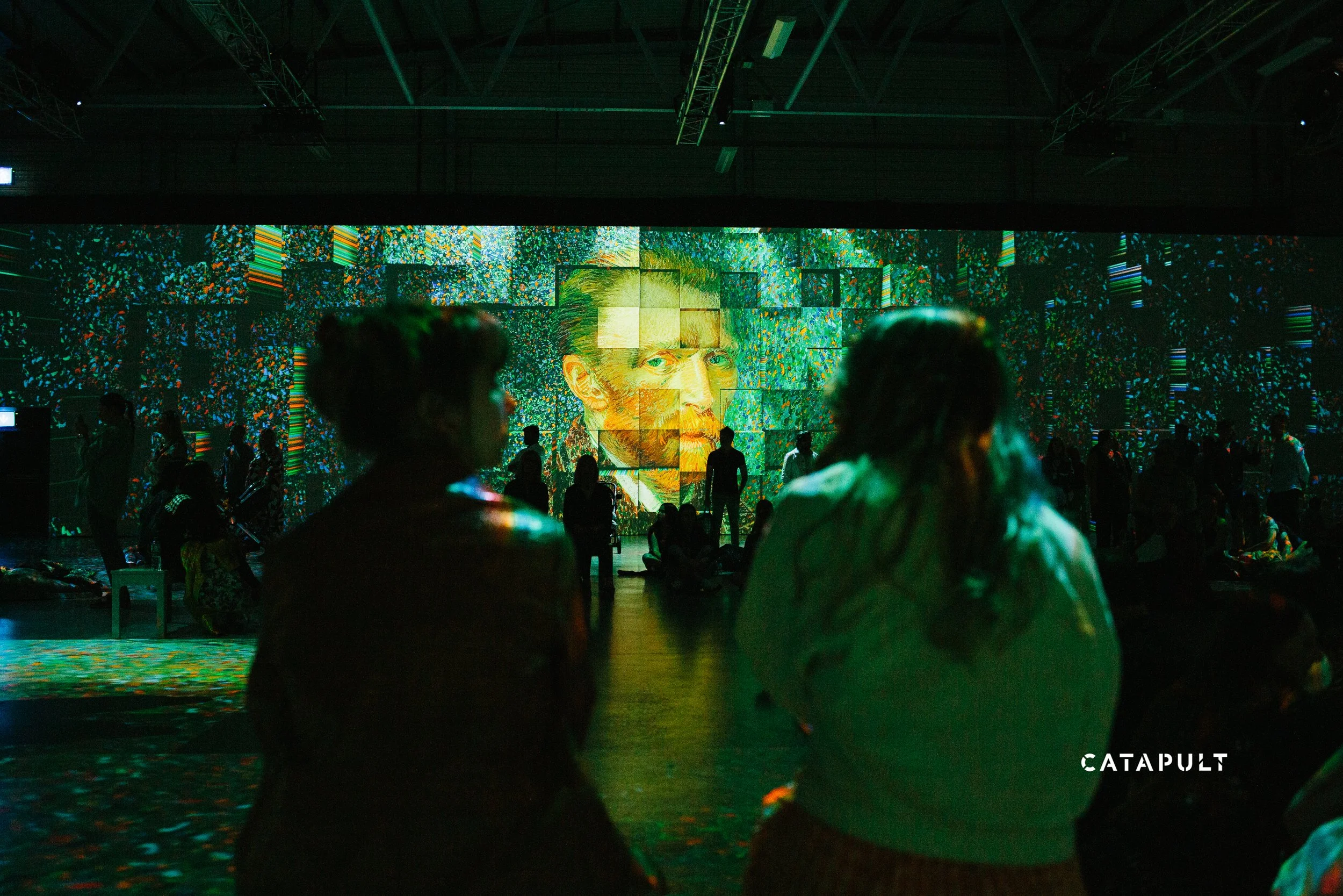 A large digital art installation displays a colorful image of Vincent van Gogh's portrait with vibrant swirling patterns. Several people are gathered in front of it, silhouetted against the display. The word 'CATAPULT' appears in the bottom right cor