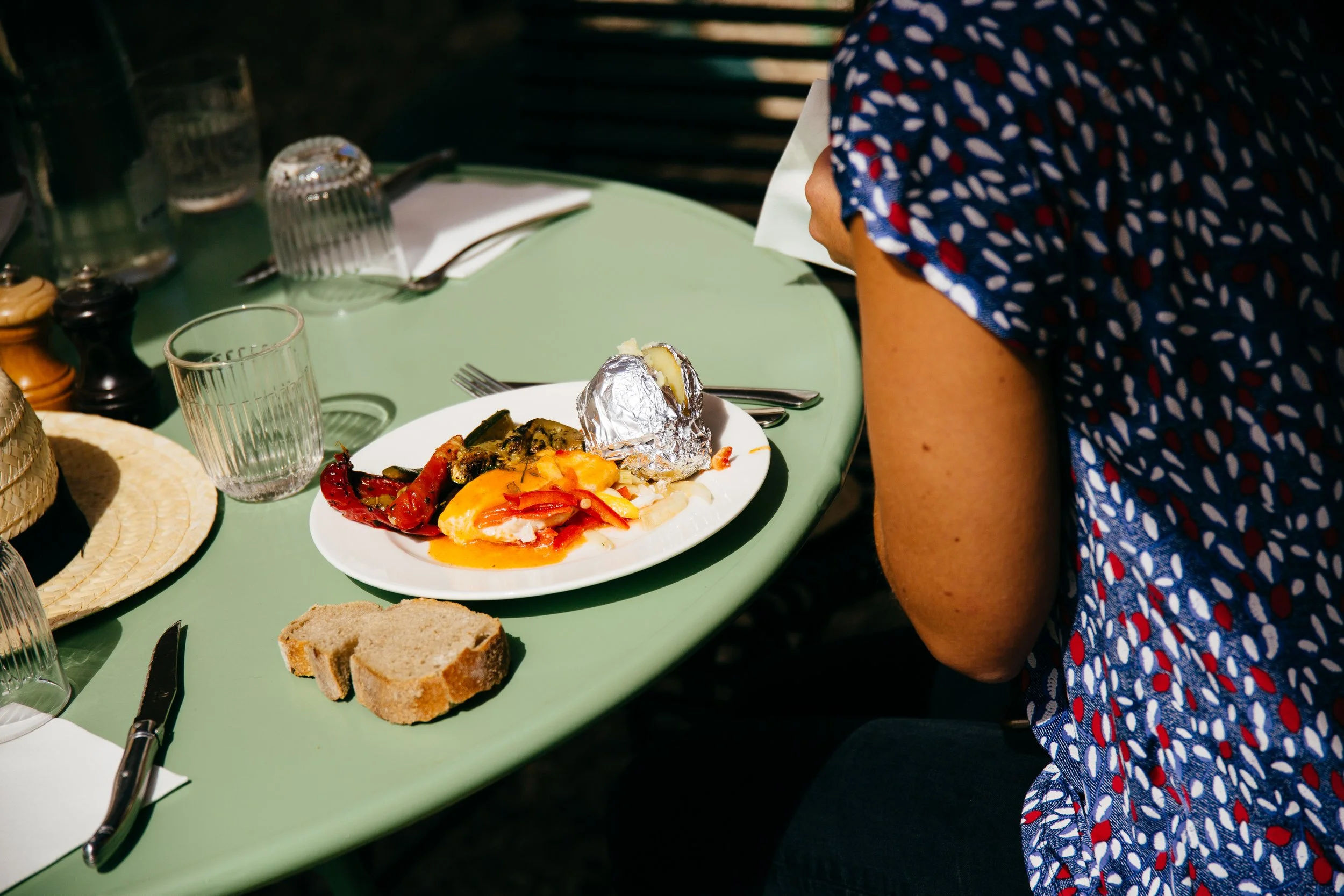 Person sitting at an outdoor table with a plate of food, including baked potato wrapped in foil, roasted vegetables, and bread slices, with glasses, a notebook, and condiments nearby.