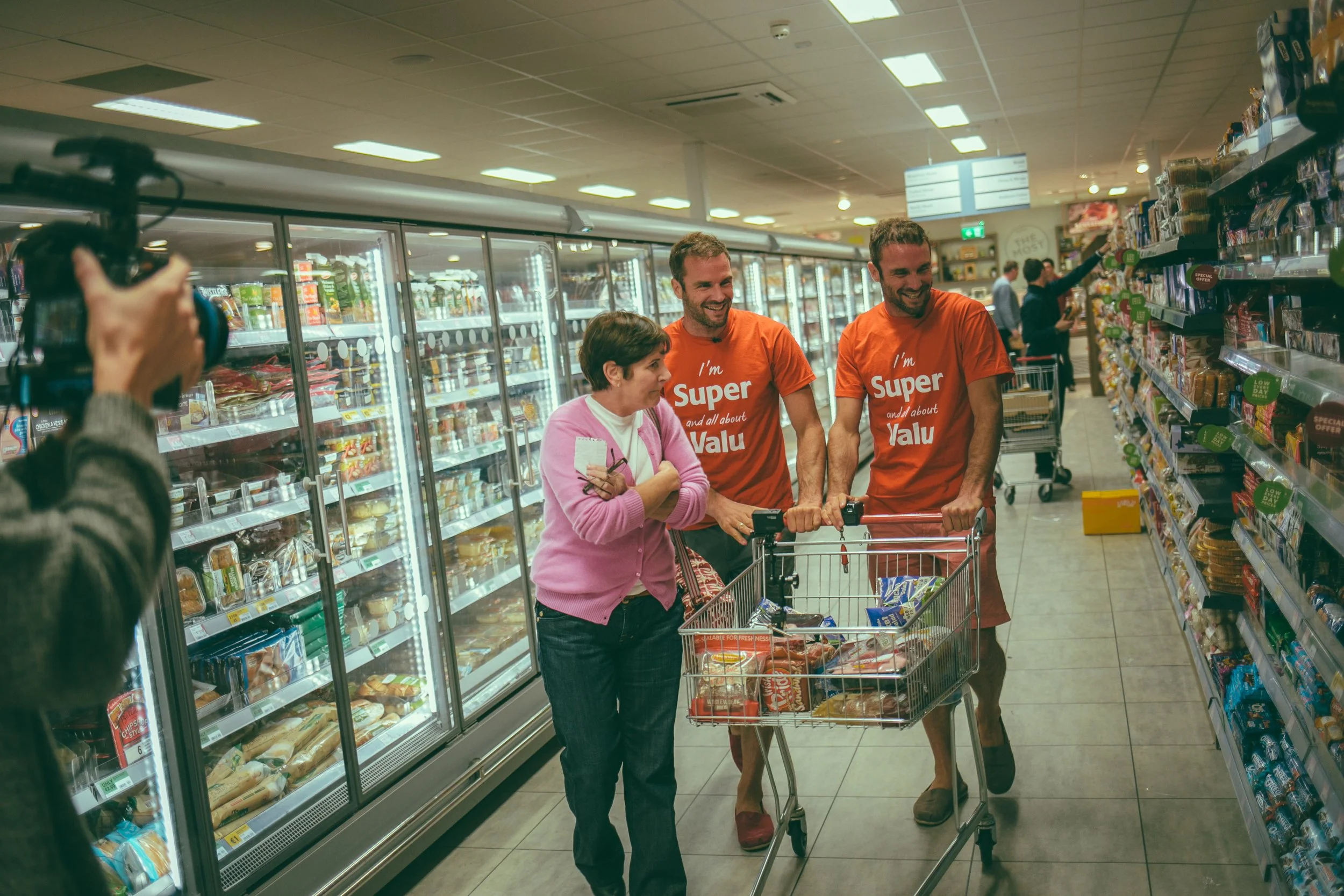 People shopping in a grocery store aisle, with two men and one woman smiling and talking while pushing a shopping cart filled with groceries.