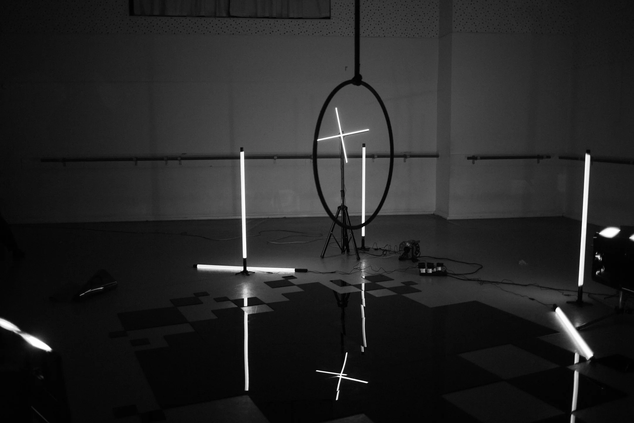 A dark room with vertical and horizontal LED light tubes, a circular LED frame, and a grid pattern on the floor, set up for an art or light installation.
