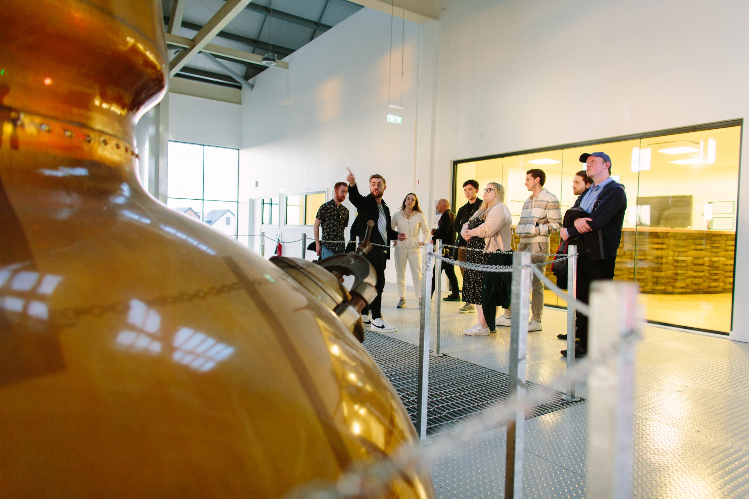 A group of people is standing behind a chain barrier inside a brightly lit industrial or museum space, with a large brass object in the foreground.