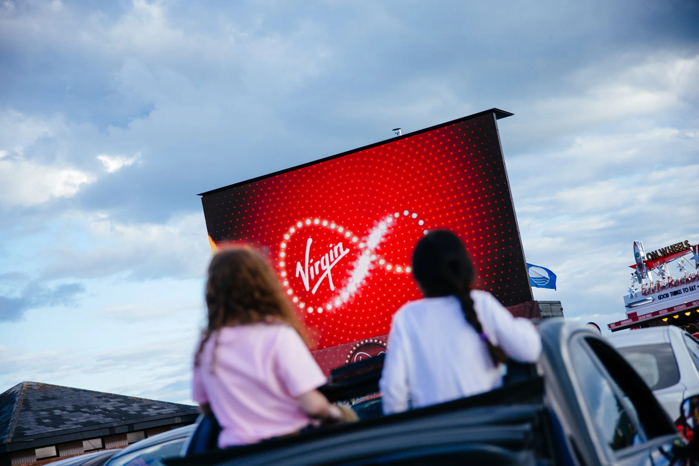 People in amusement park rides watching a large red screen displaying a Virgin logo in a cloudy sky.