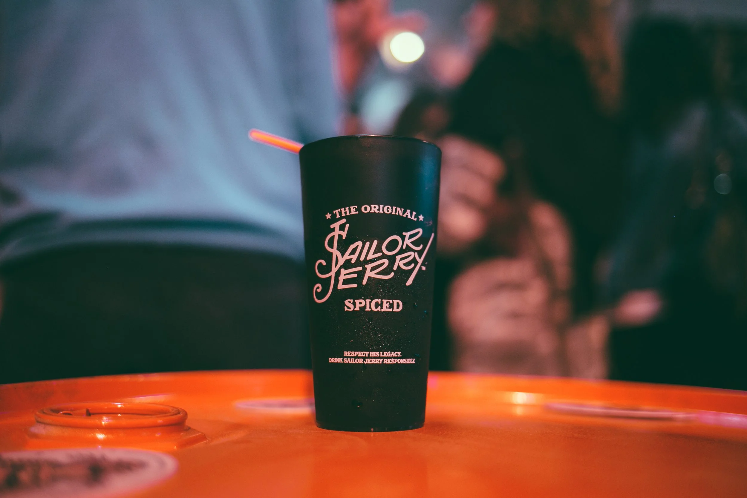 Close-up of a black travel mug with label reading 'The Original Sailor Jerry Spiced' sitting on an orange surface, with a blurred background and a person's hand and wrist visible holding a cigarette.
