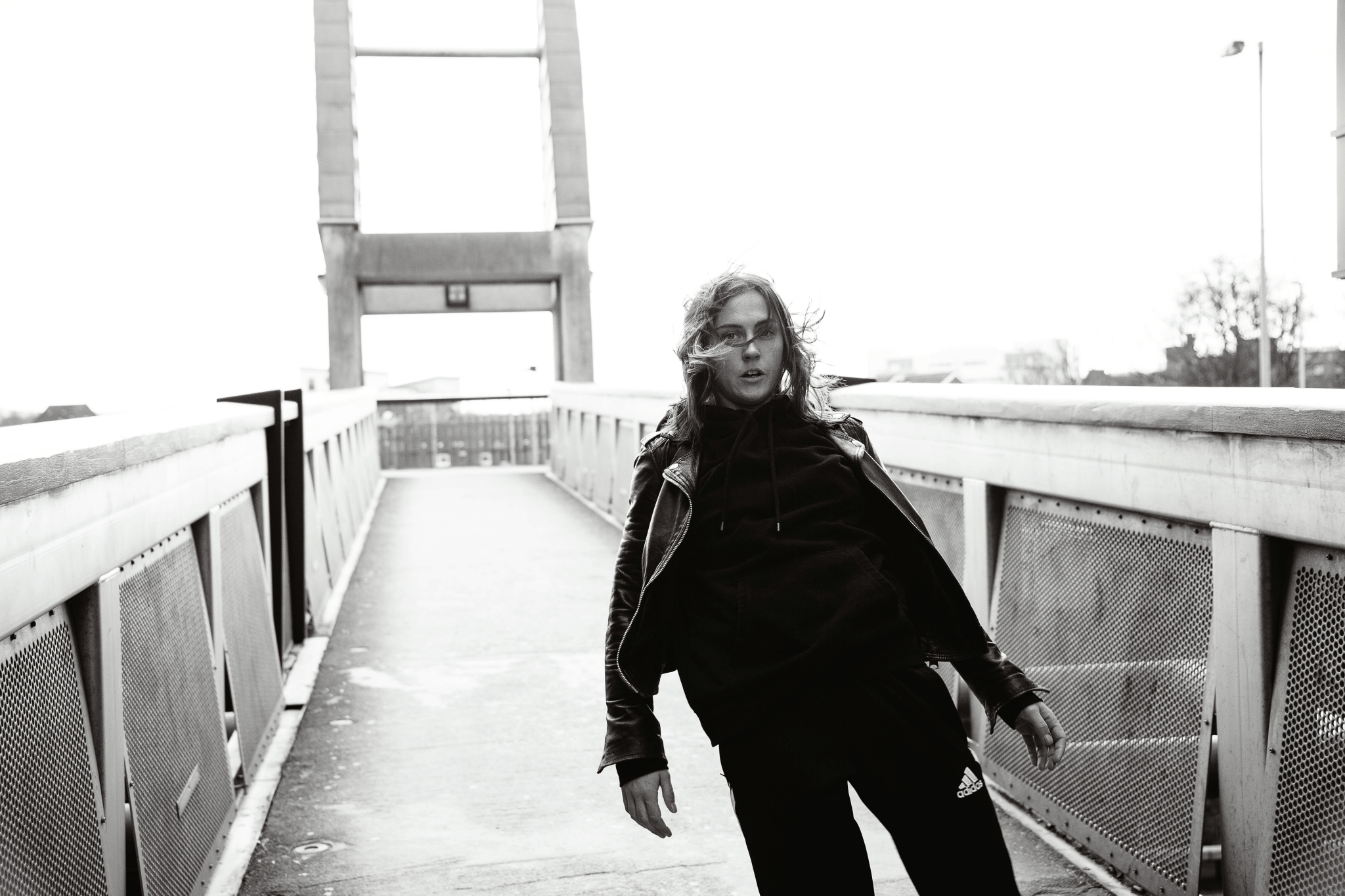 Black and white photo of a young woman with glasses and long wavy hair, wearing a black hoodie and leather jacket, leaning against a railing on a pedestrian bridge with an overcast sky.