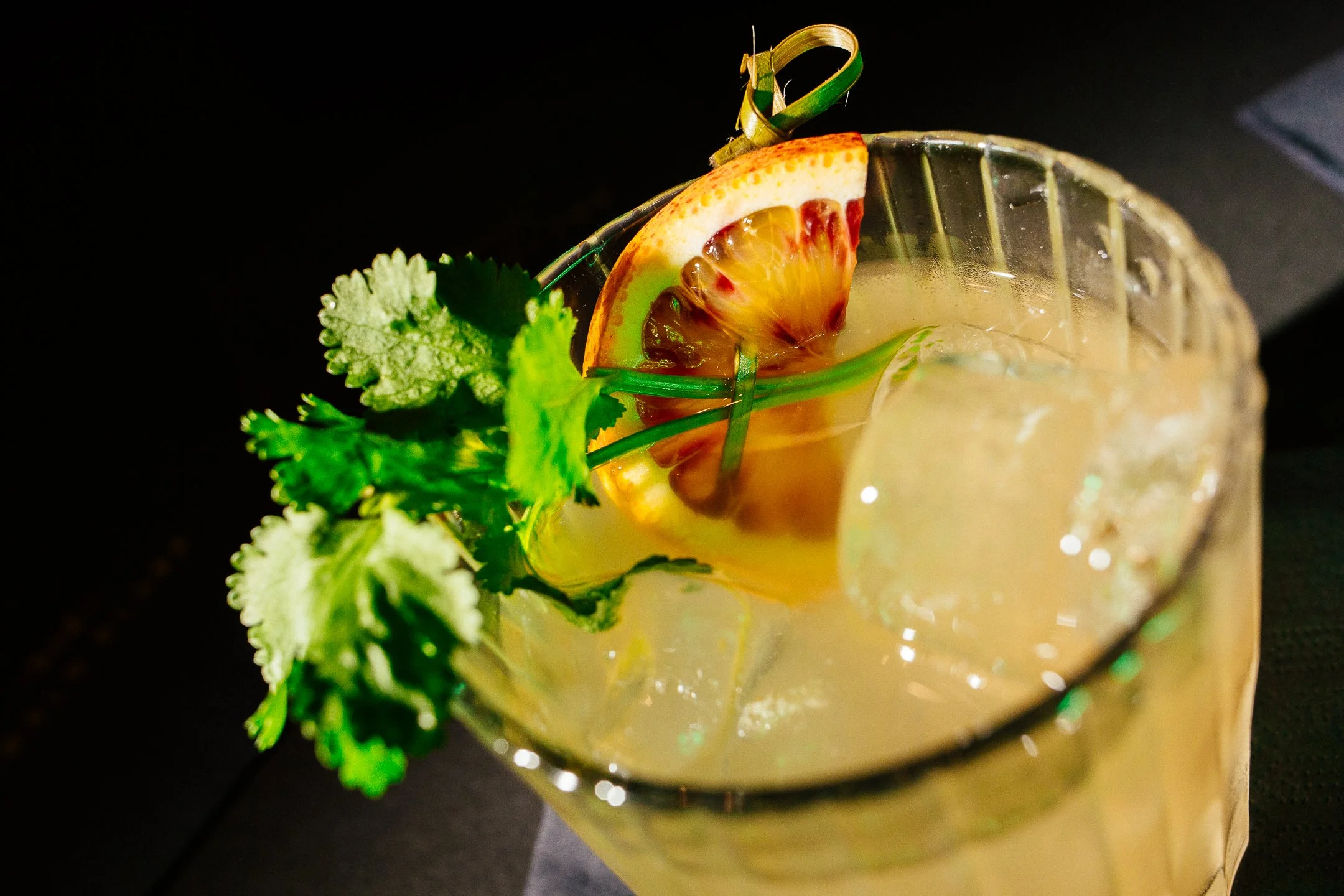 A cocktail garnished with a slice of orange, lime, and a sprig of cilantro, served in a glass with ice.