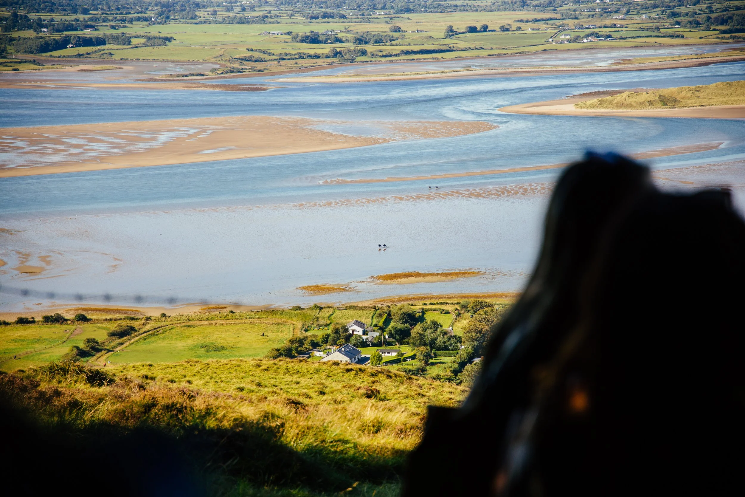A scenic view of a landscape with a river or estuary, sandy banks, green grassy fields, and scattered houses in the background. The photo appears to be taken from a higher vantage point, possibly from a hill or mountain, with part of a person's dark 