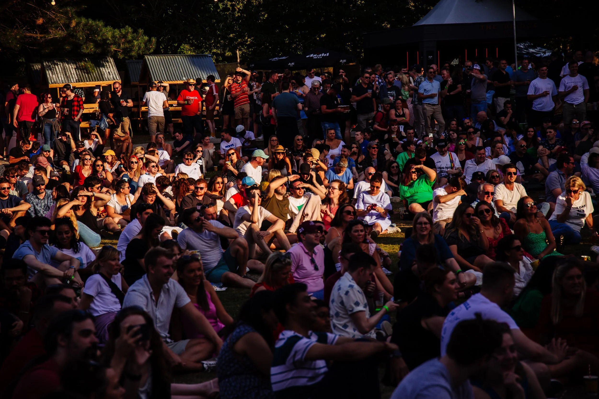 A large crowd of people gathered outdoors sitting on the ground and standing, likely watching a performance or event, with some holding drinks, some taking photos, and enjoying themselves, under trees on a bright day.
