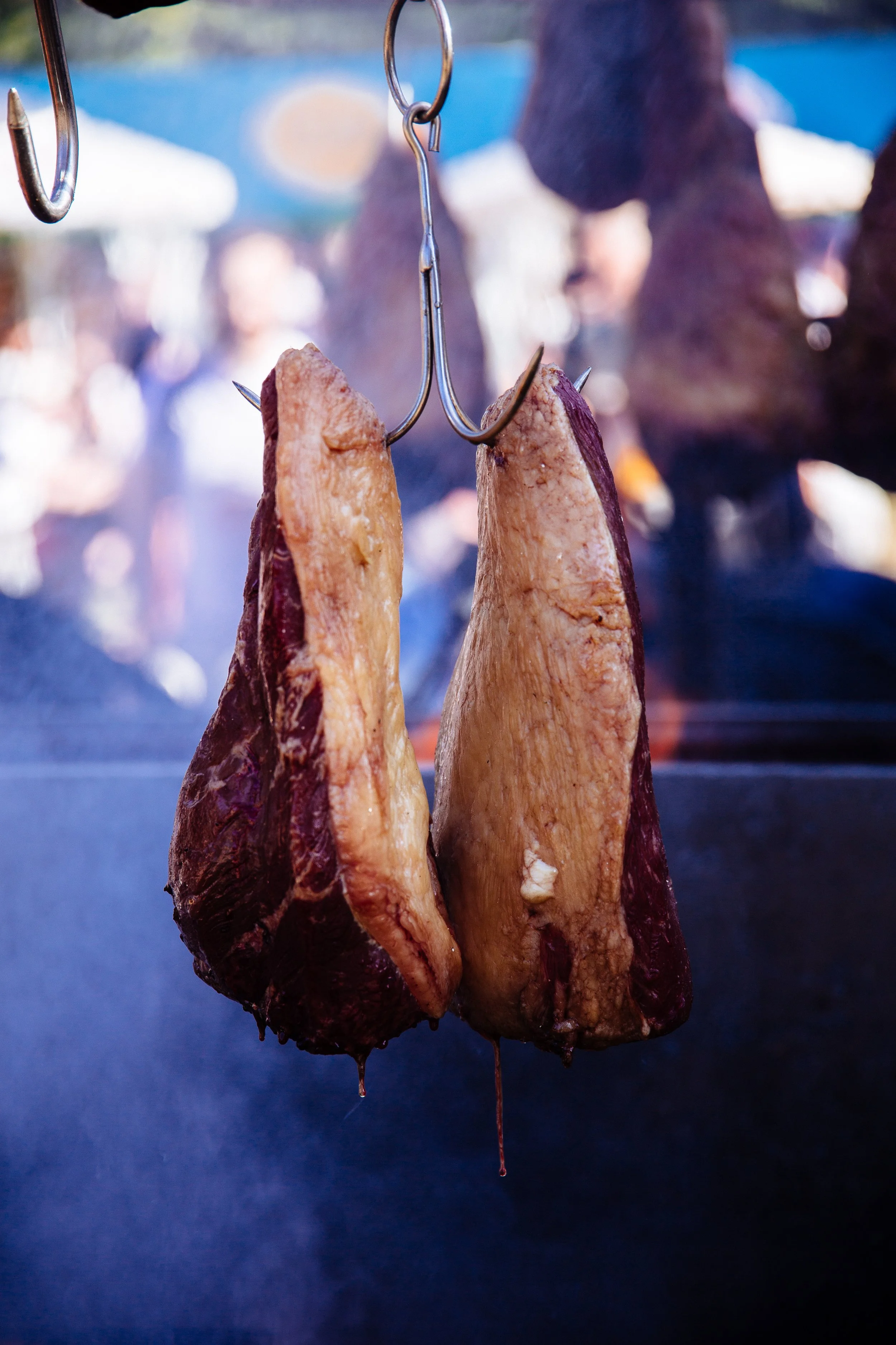 Hanging cooked pork belly strips with dripping juices in front of a blurred background at a market or food stall.