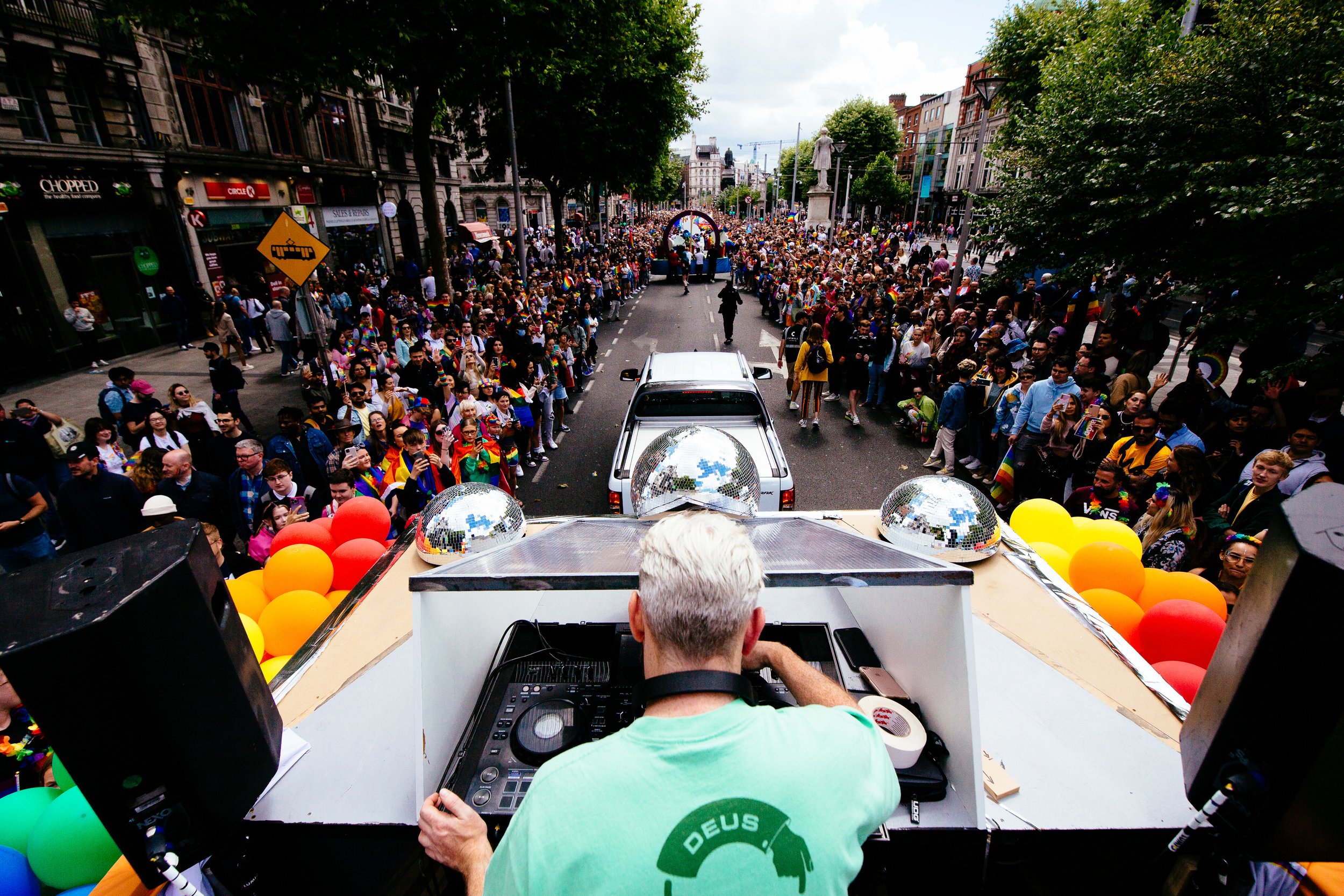 A DJ is playing music on a sound system inside a parade float decorated with multicolored balloons, as a large crowd of people gathers along the street for a pride parade.