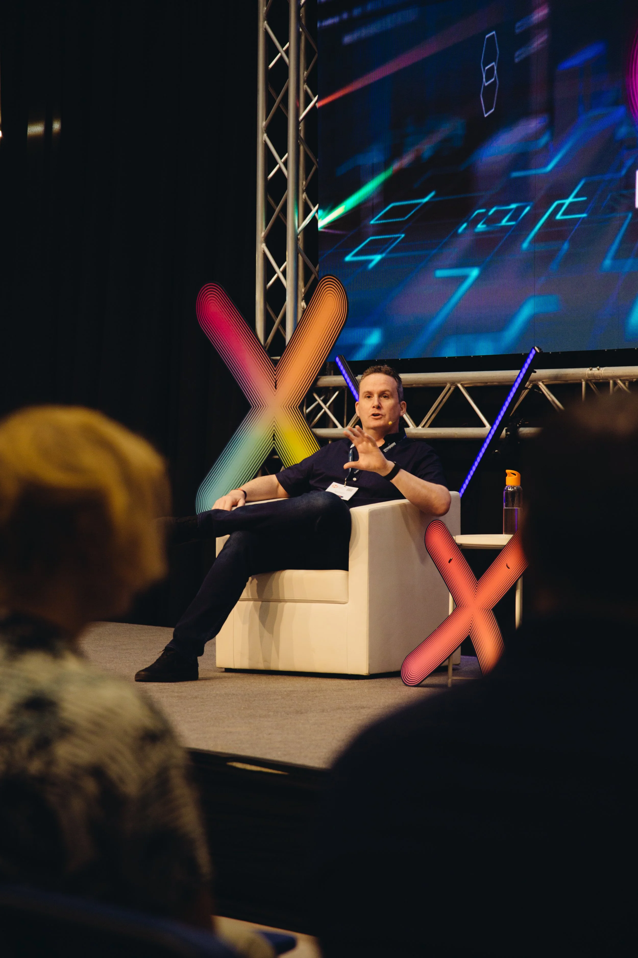 A man is sitting on a white armchair on stage, gesturing with his hand while speaking at a conference. Behind him, there is a large digital screen displaying a futuristic, circuit-like graphic. On the left side of the stage, there is a colorful illum