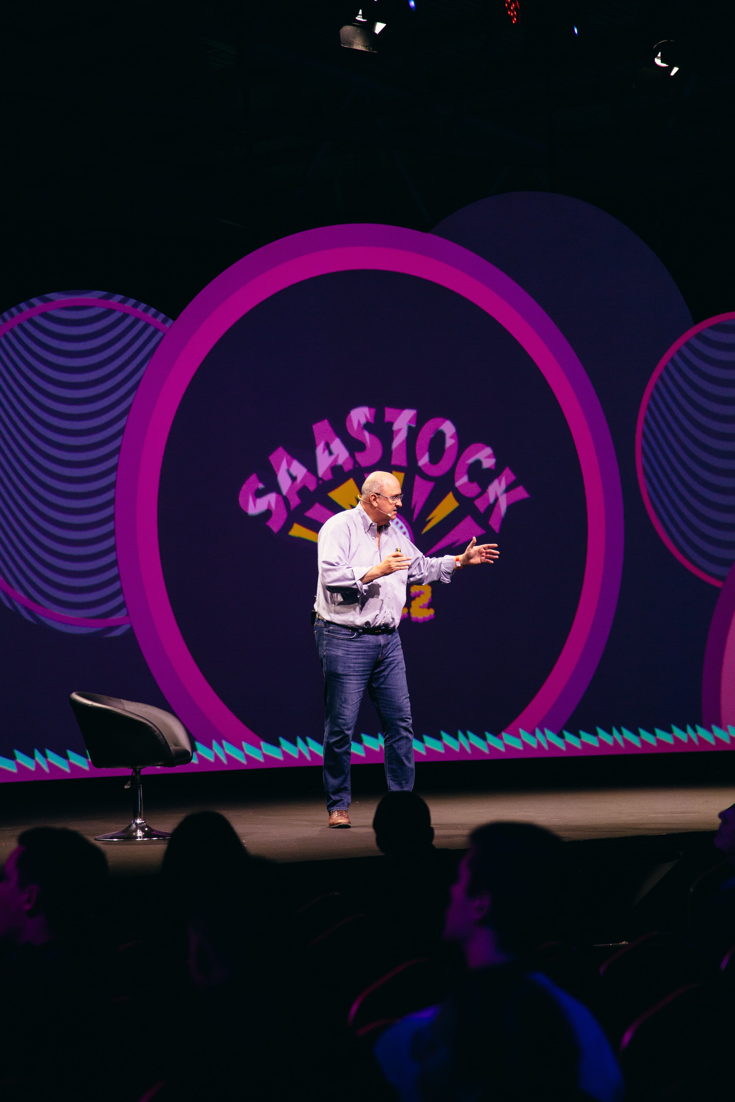 A man with glasses, bald head, wearing a striped shirt and jeans, talks on a stage with a microphone in hand. Behind him is a large screen with vibrant pink, purple, and blue graphics, including the text "SaaS Touch" and the number "2". There is a bl
