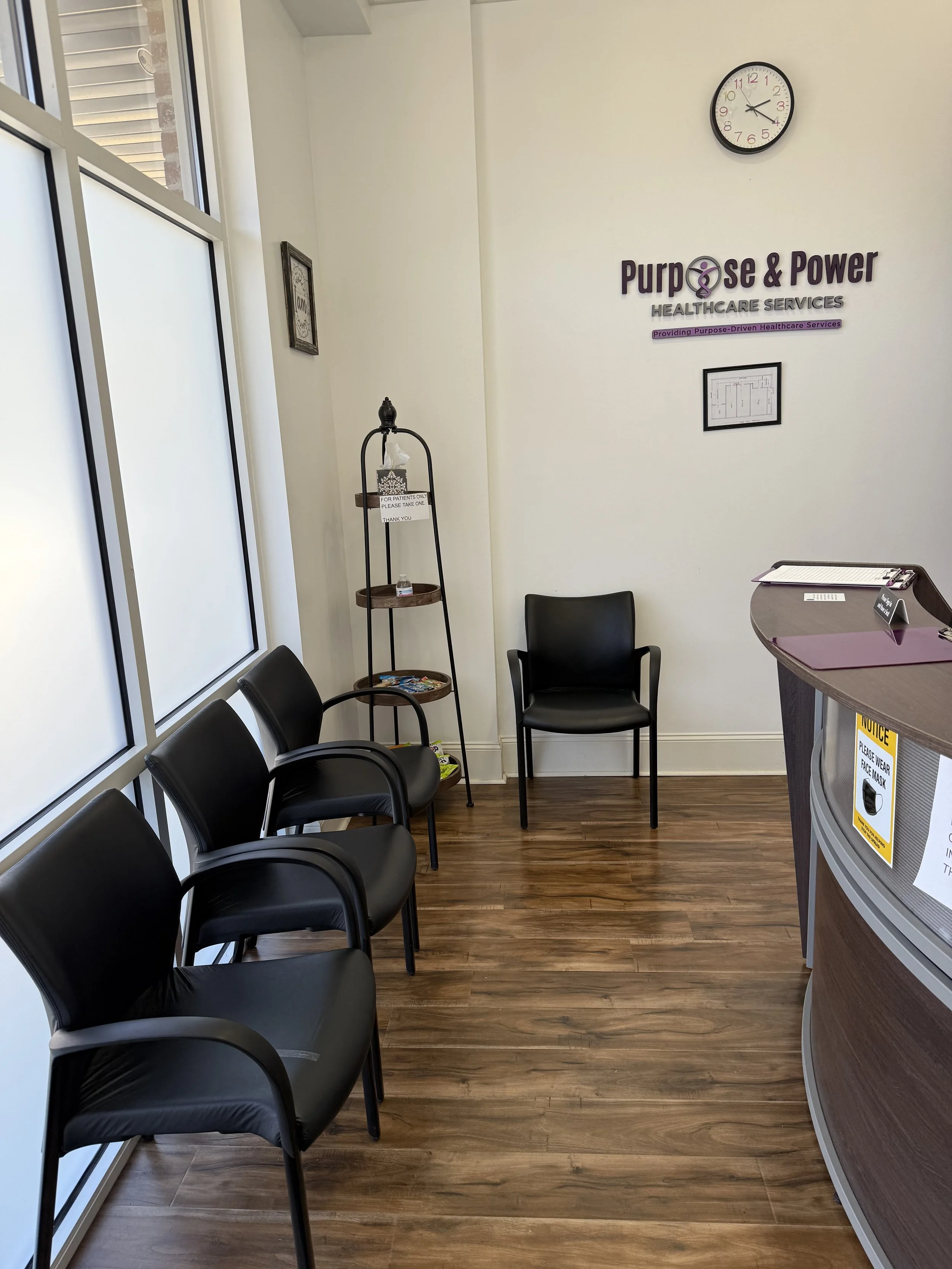 Waiting room at Purpose & Power Healthcare Services with five black chairs, a small wooden shelf with hand sanitizer and magazines, a reception desk with notices, a clock on the wall showing 2:15, and a directory map.