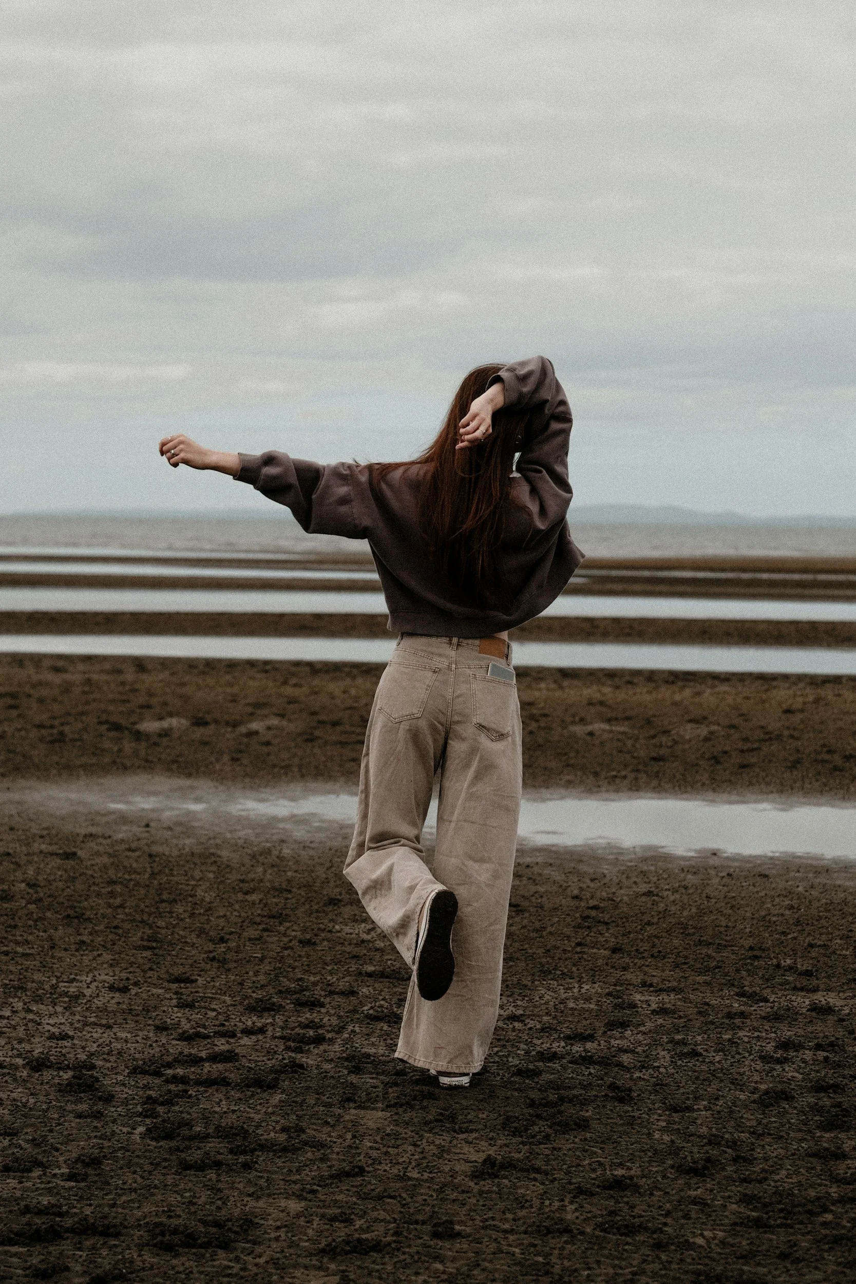 A person with long hair wearing a dark oversized sweatshirt and beige pants is balancing on one foot in a muddy outdoor area, with a cloudy sky and water in the background.