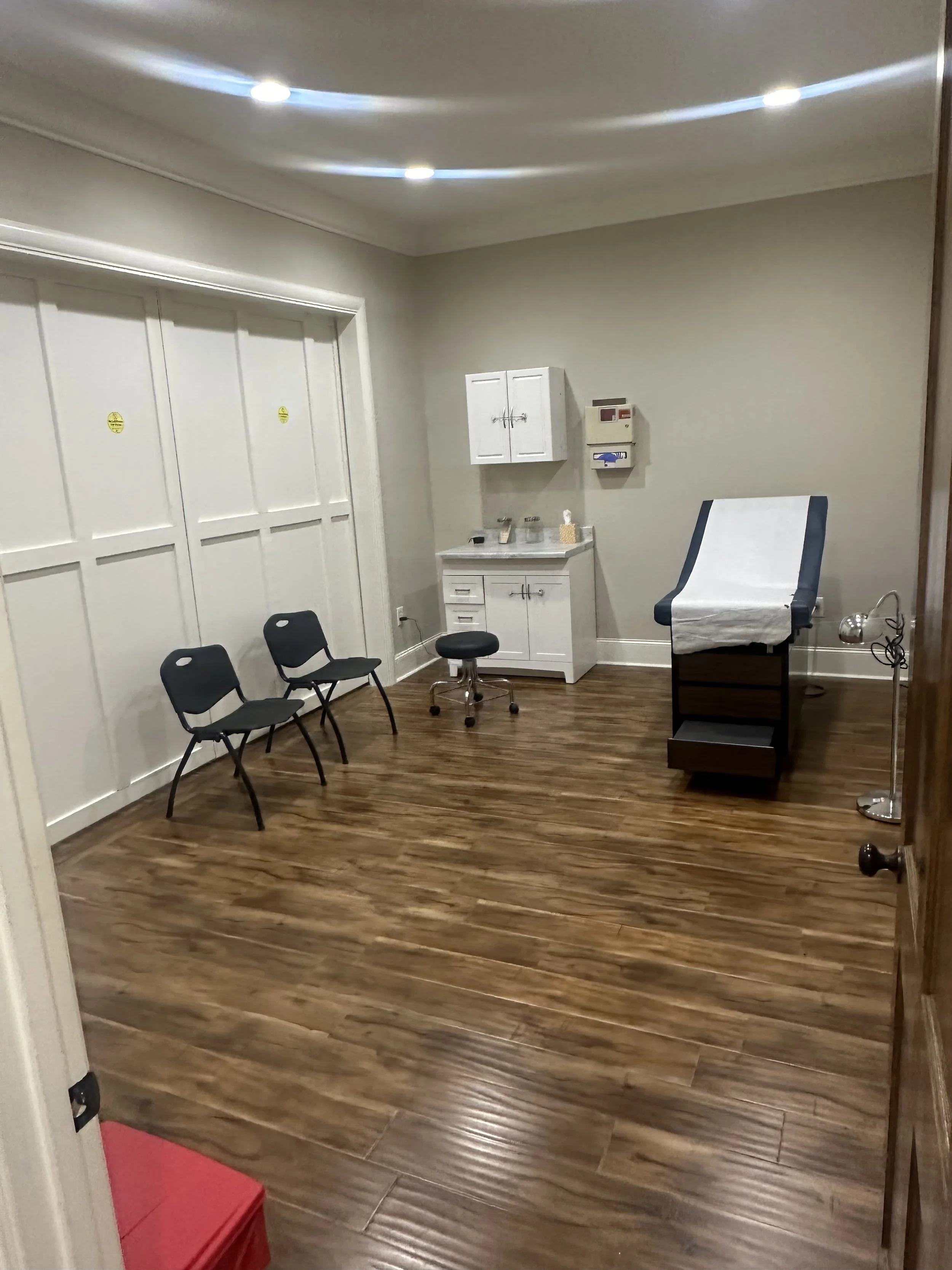 Room in a medical facility with wooden floors, white walls, a white cabinet, sterile supplies, and examination table covered with paper.