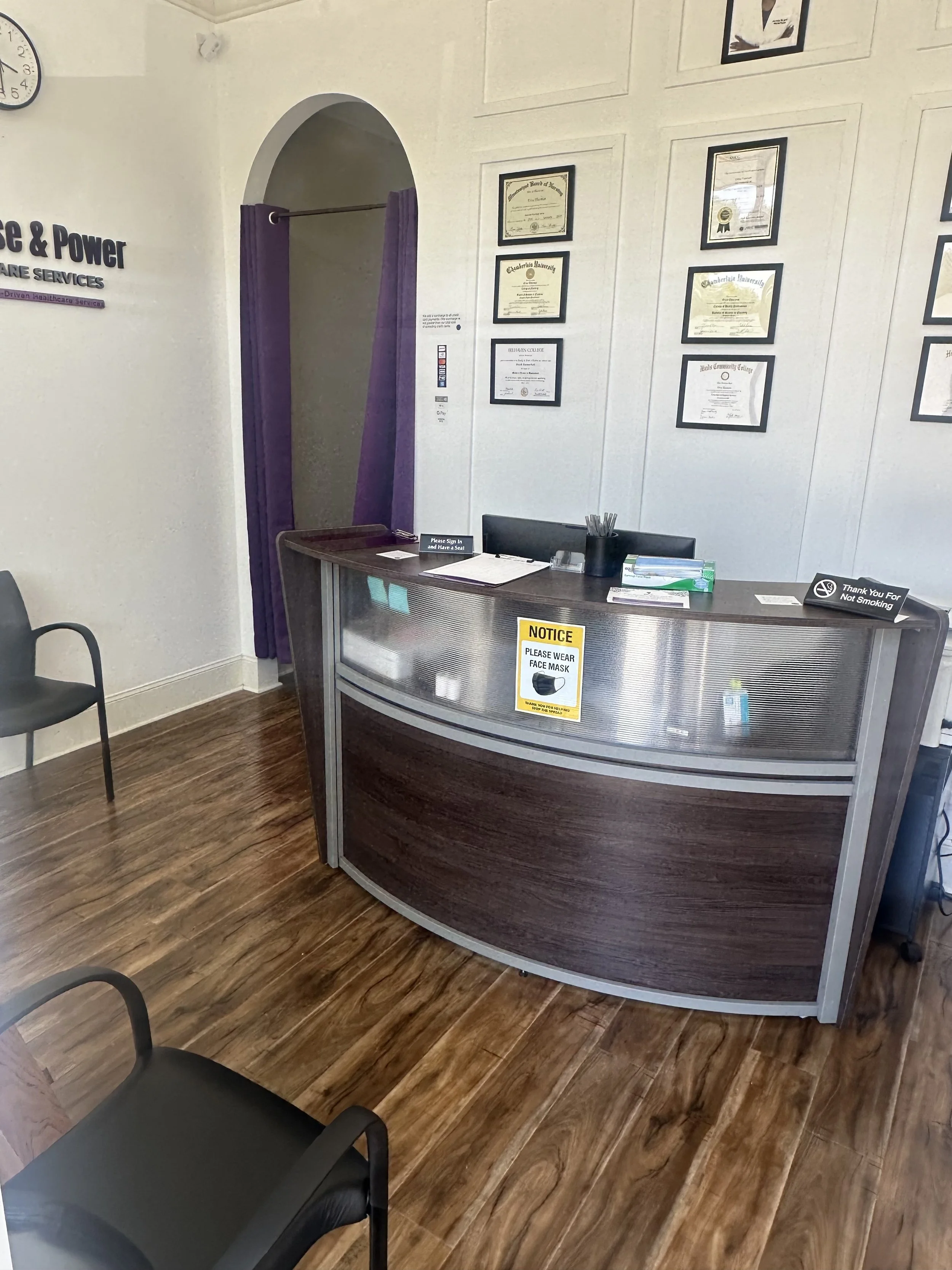 Reception desk in an office or clinic with framed certificates on the wall, a sign requesting face masks, and chairs nearby.