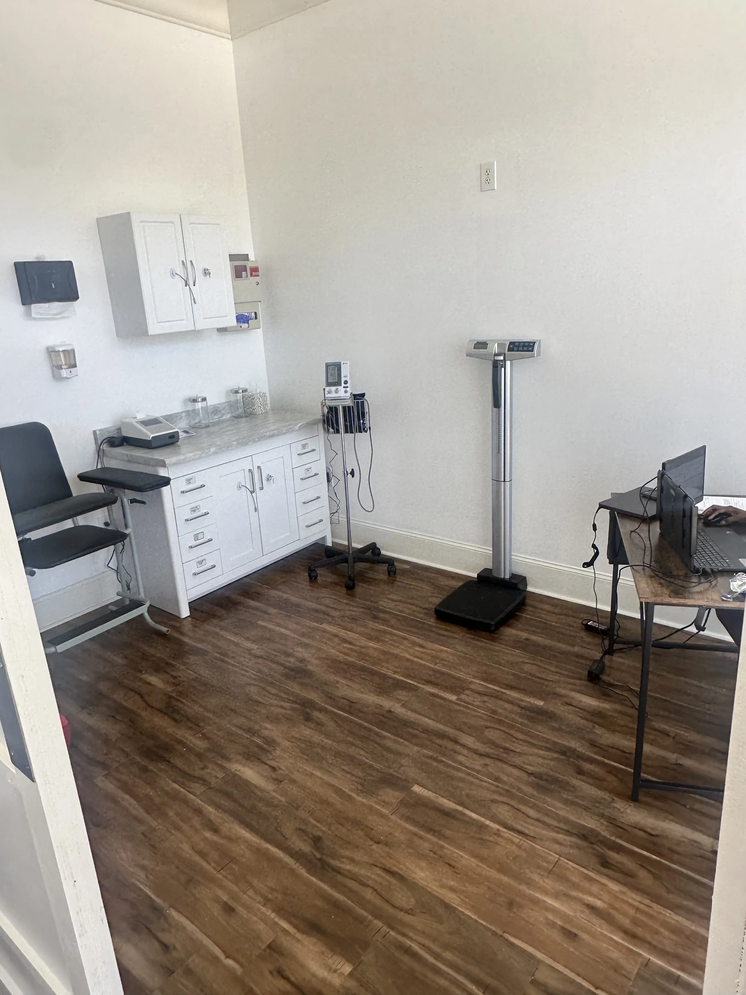 Empty medical examination room with a white cabinet, a scale, and a computer on a small table.