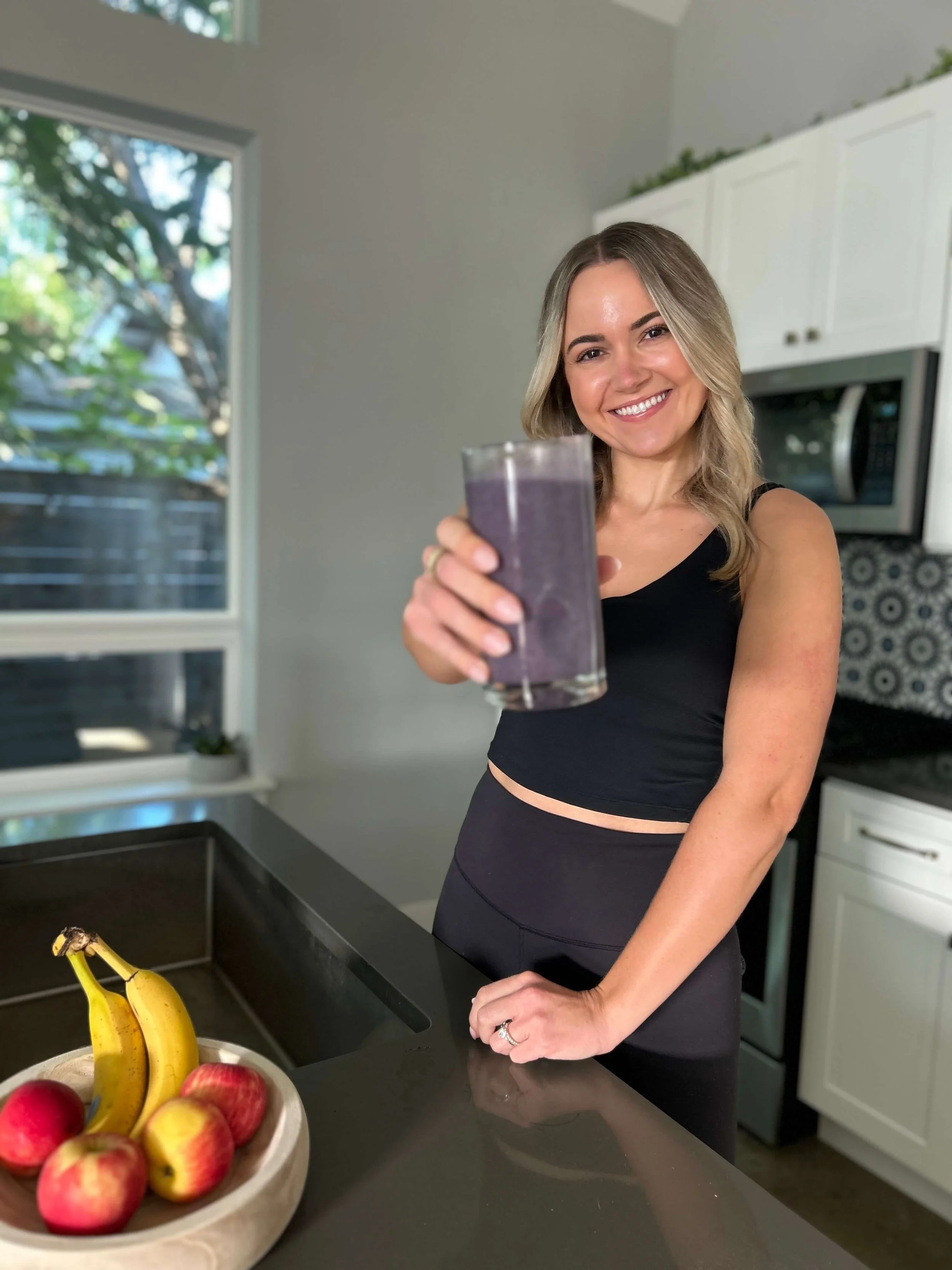 woman holding a glass with a blue smoothie, fruit sitting in front of her in kitchen