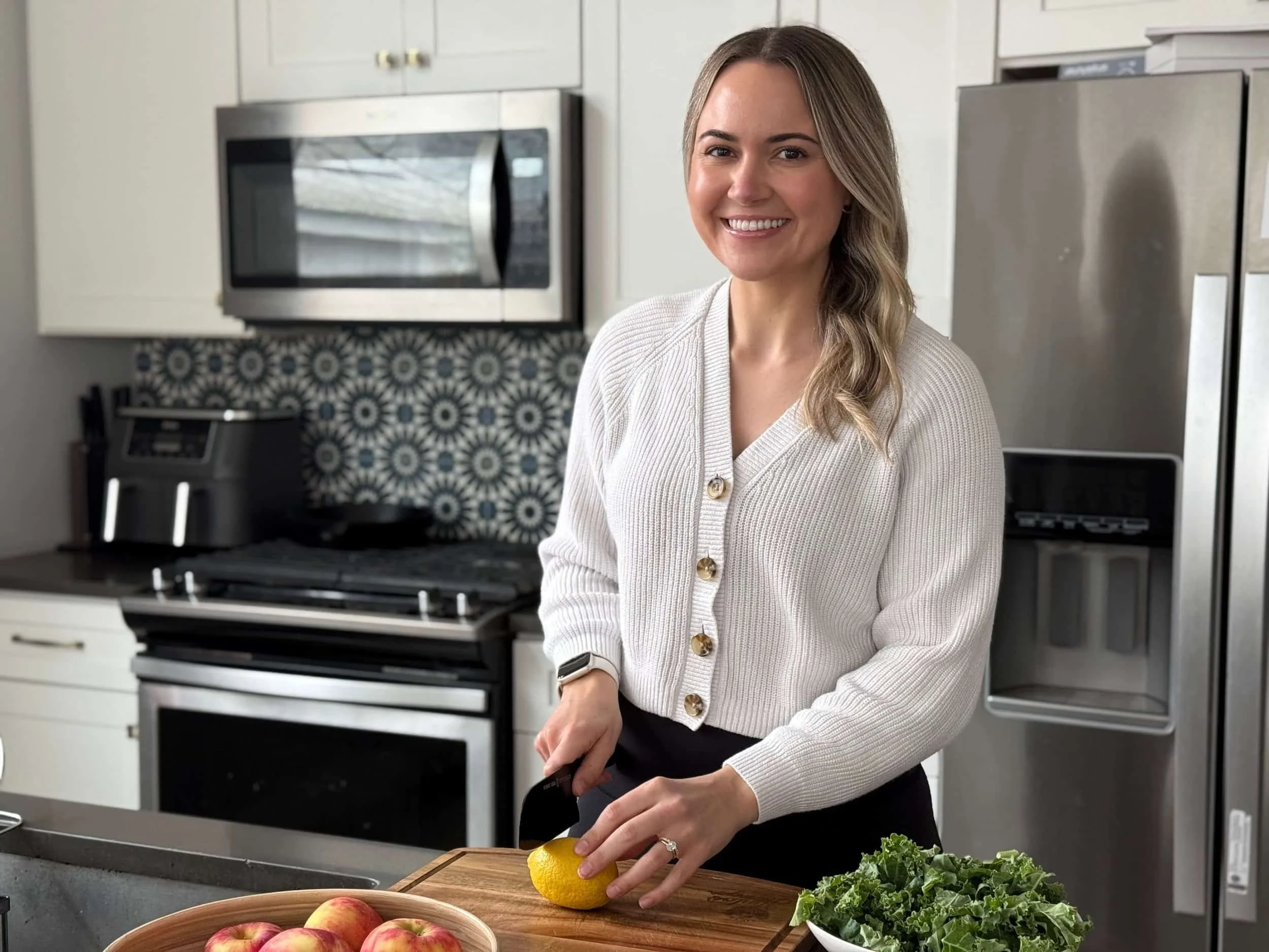 A woman with blonde hair, smiling, peeling a lemon in a modern kitchen with white cabinets, stainless steel appliances, and a patterned black and white backsplash.