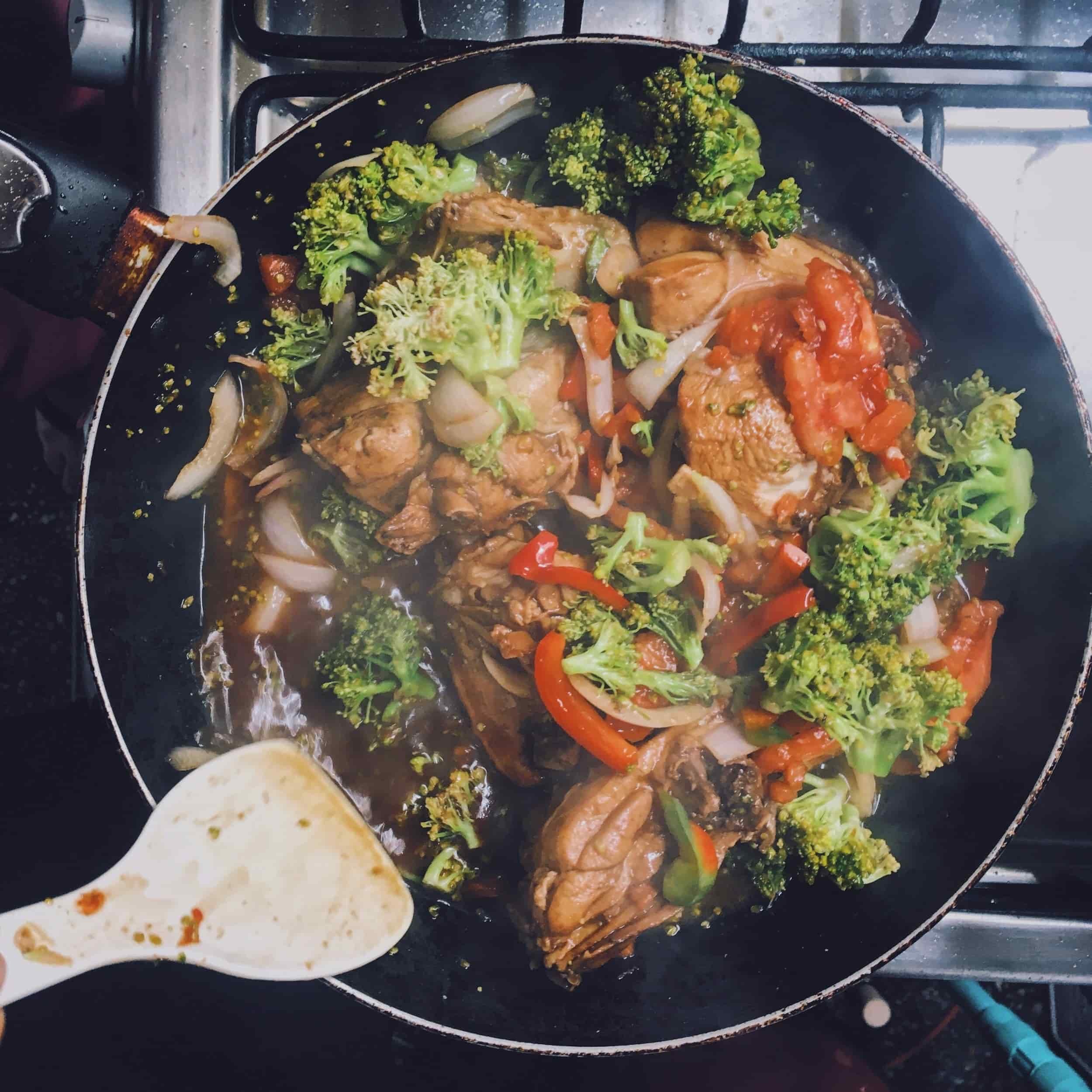 Cooking stew with chicken, broccoli, onions, and tomatoes in a black skillet on a stove.