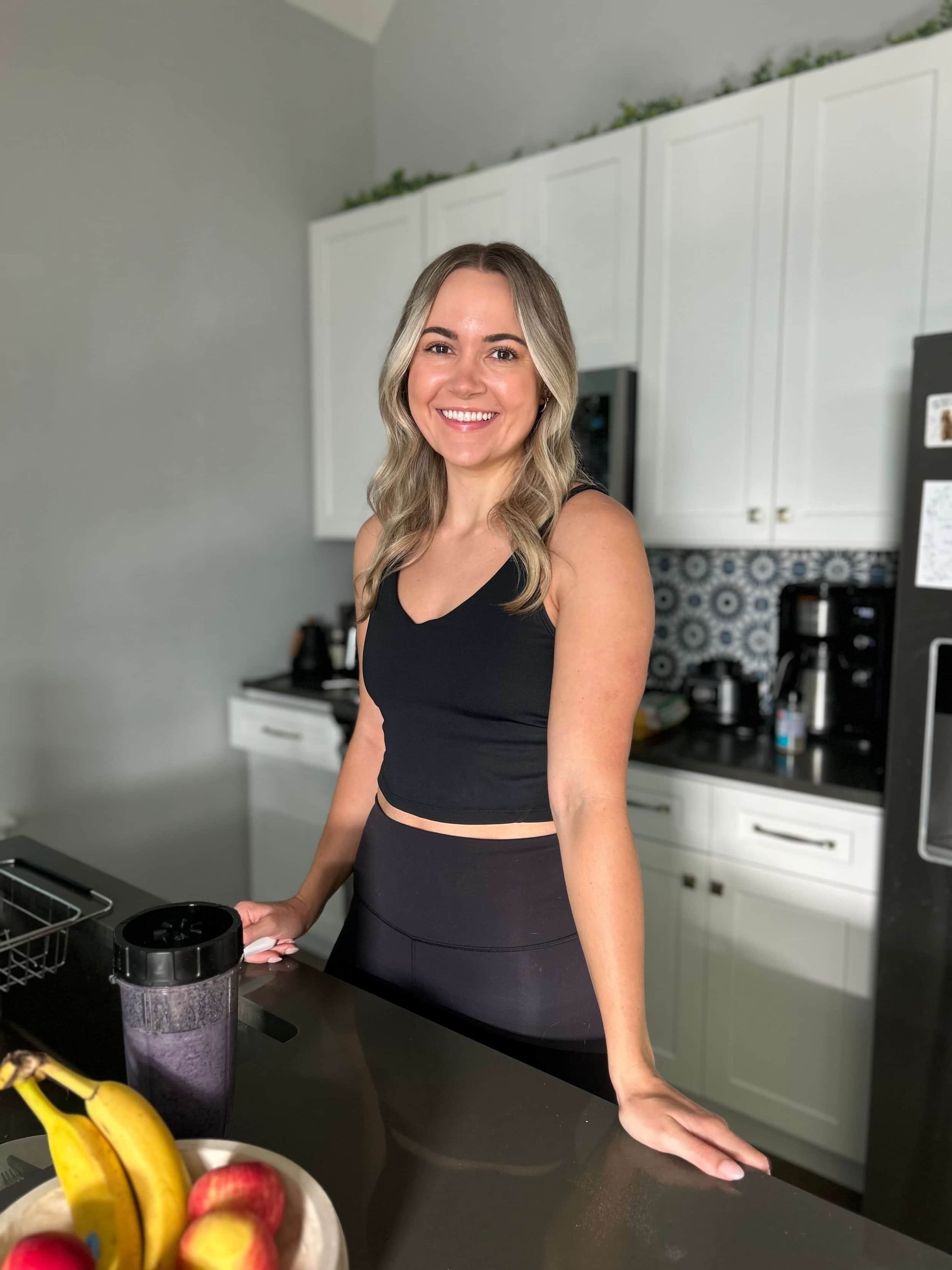 A woman with long, wavy blonde hair, wearing a black sleeveless top and black leggings, smiling in a kitchen with white cabinets, a patterned backsplash, and a black countertop, holding a blender bottle.