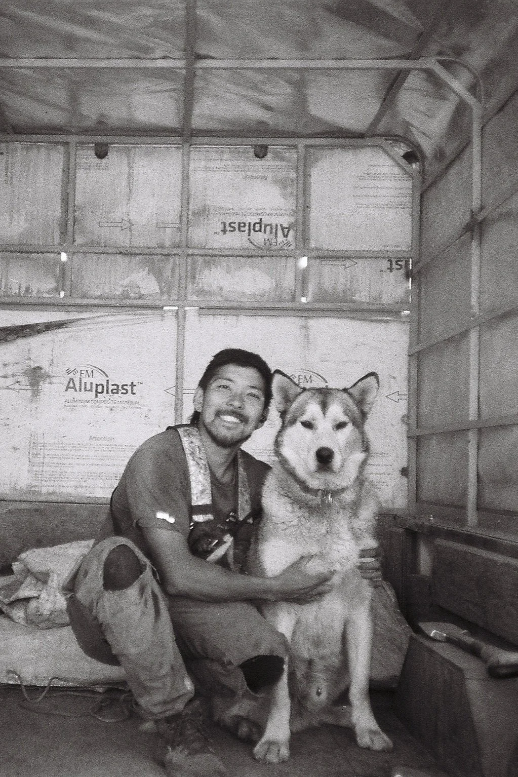 A man sitting on the floor inside a truck with a husky dog, smiling at the camera.