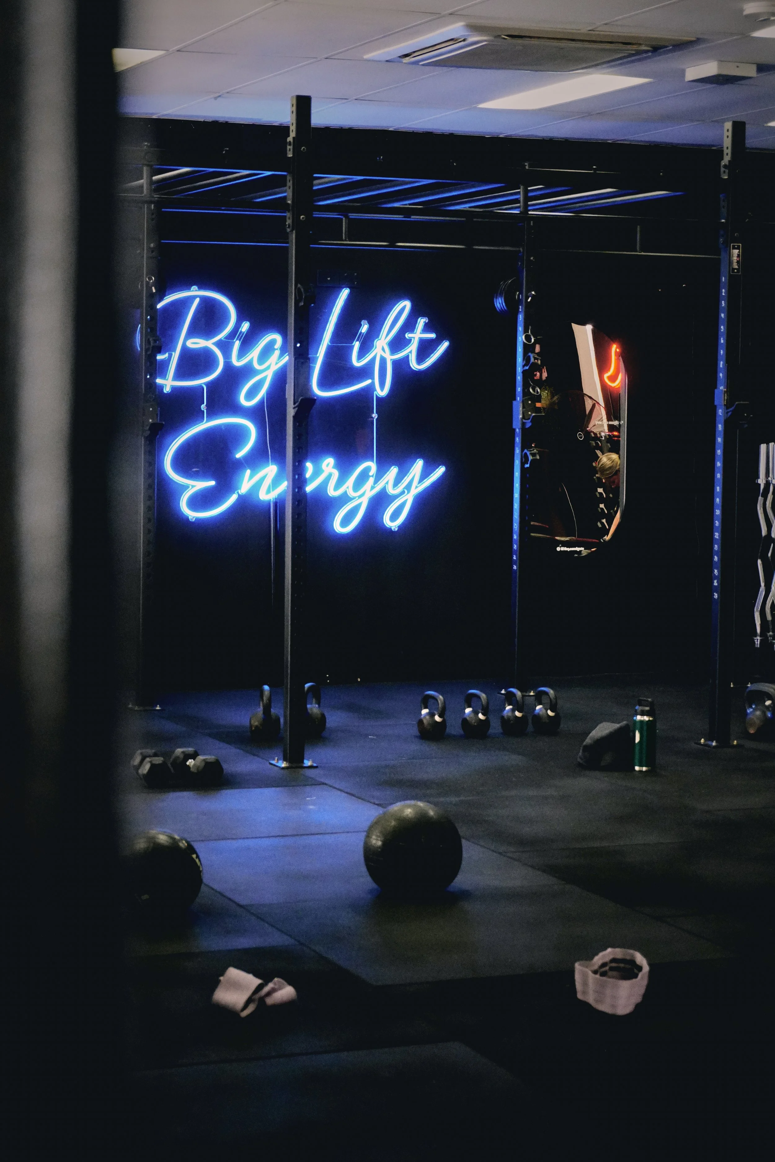 A gym with a neon sign that reads 'Big Lift Energy' on a black wall. There are kettlebells on the floor, workout gloves, and a mirror reflecting gym equipment and a person wearing a hat.