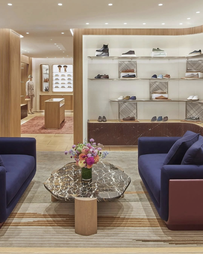 A shoe display store with shelves of shoes, a mannequin dressed in a patterned outfit, and a seating area with two navy blue sofas and a marble coffee table with a flower arrangement.