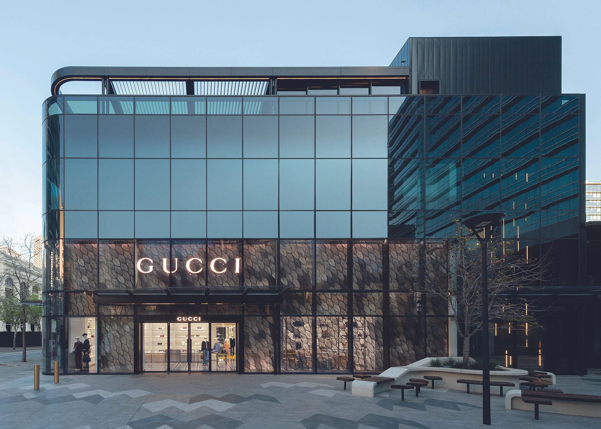Modern glass building with Gucci store on the ground floor, people entering and walking outside, trees and benches in the surrounding area.