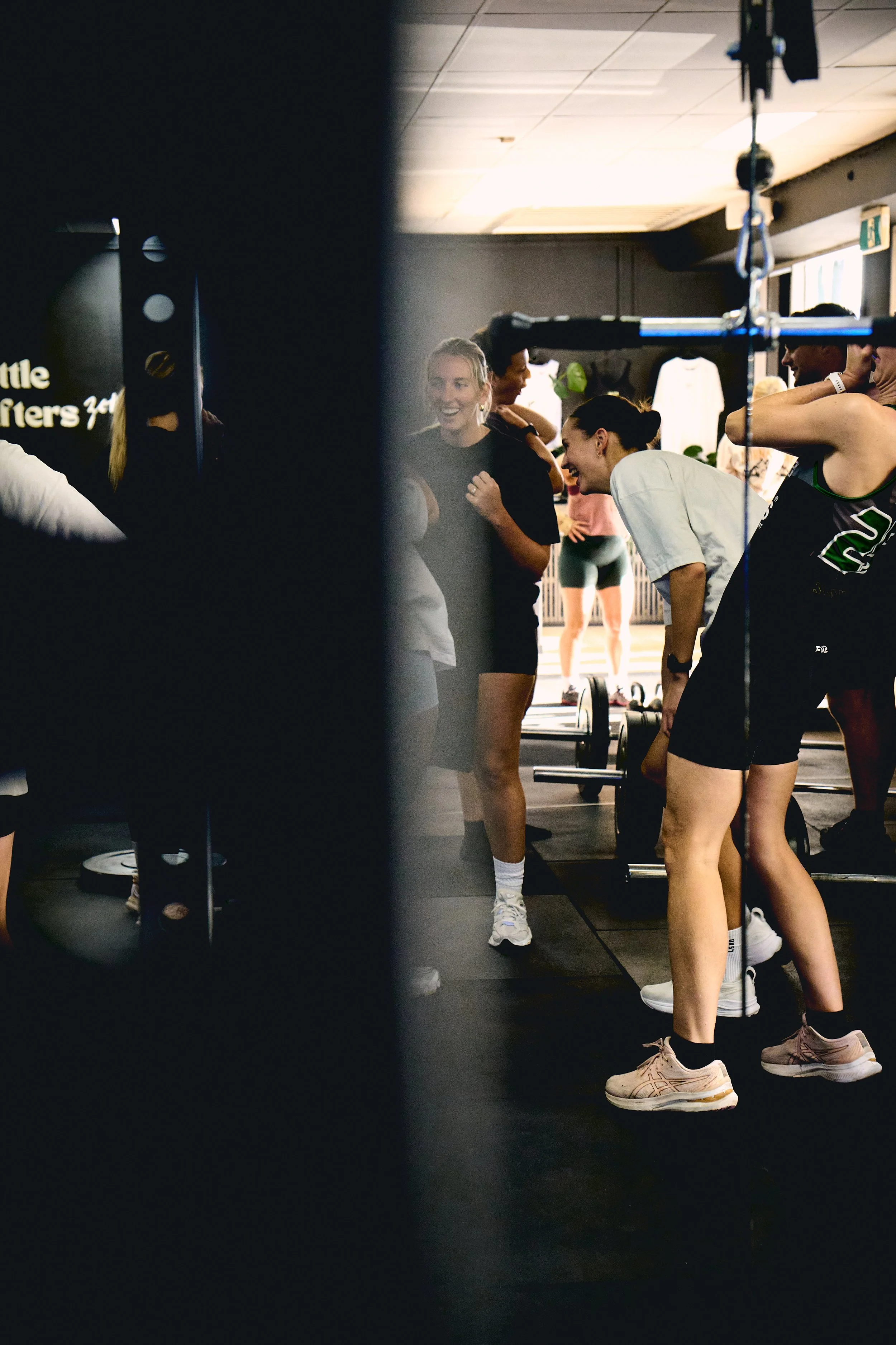 People at a gym, engaging in a fitness class or workout session, with some smiling and interacting, surrounded by gym equipment like dumbbells and workout gear.