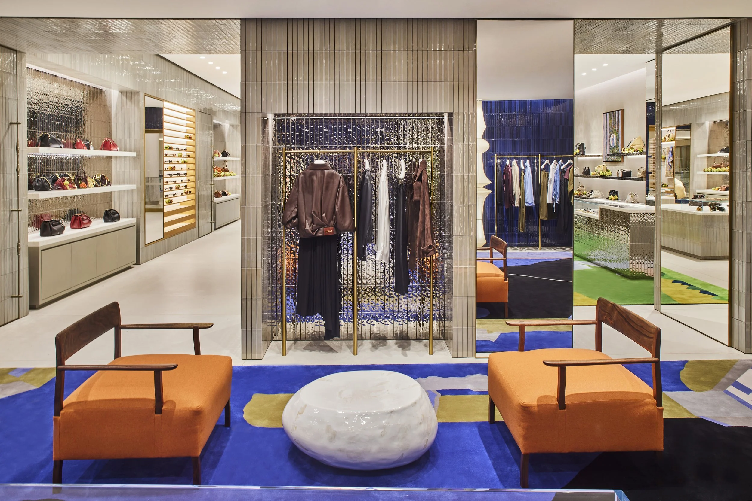 Interior of a modern clothing store displaying handbags, clothes, and accessories with seating area featuring orange chairs and a white marble table.