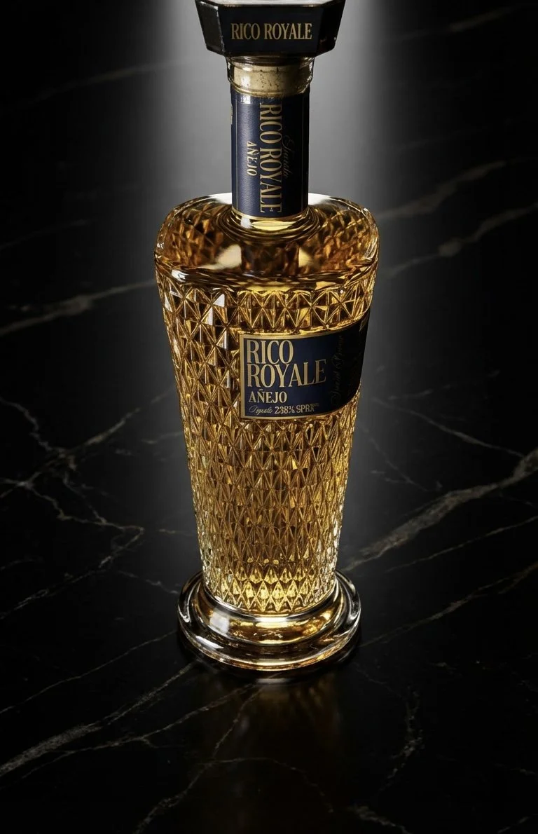 A decorative glass bottle of Rico Royale Añejo tequila with a dark label, sitting on a polished black marble surface.