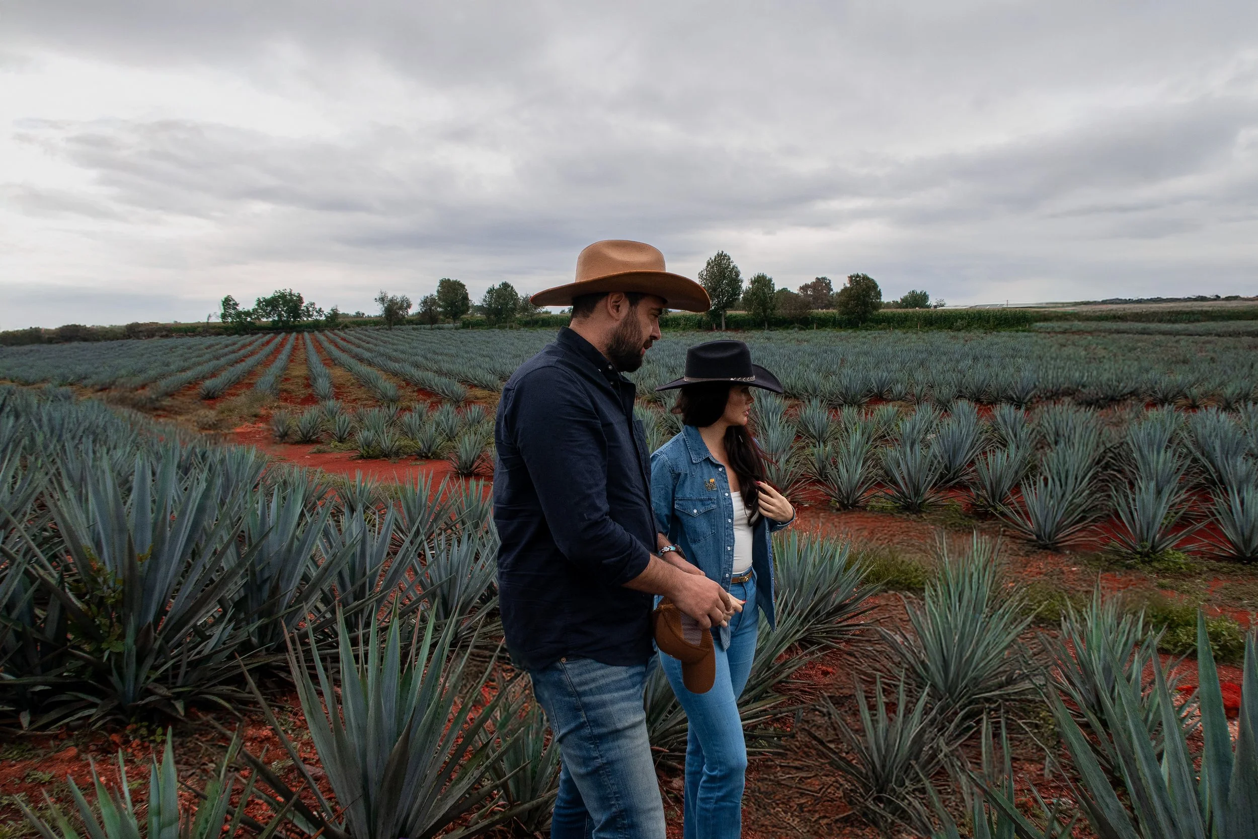 A man and woman walking through an agave field on a cloudy day, wearing cowboy hats and casual denim clothing.