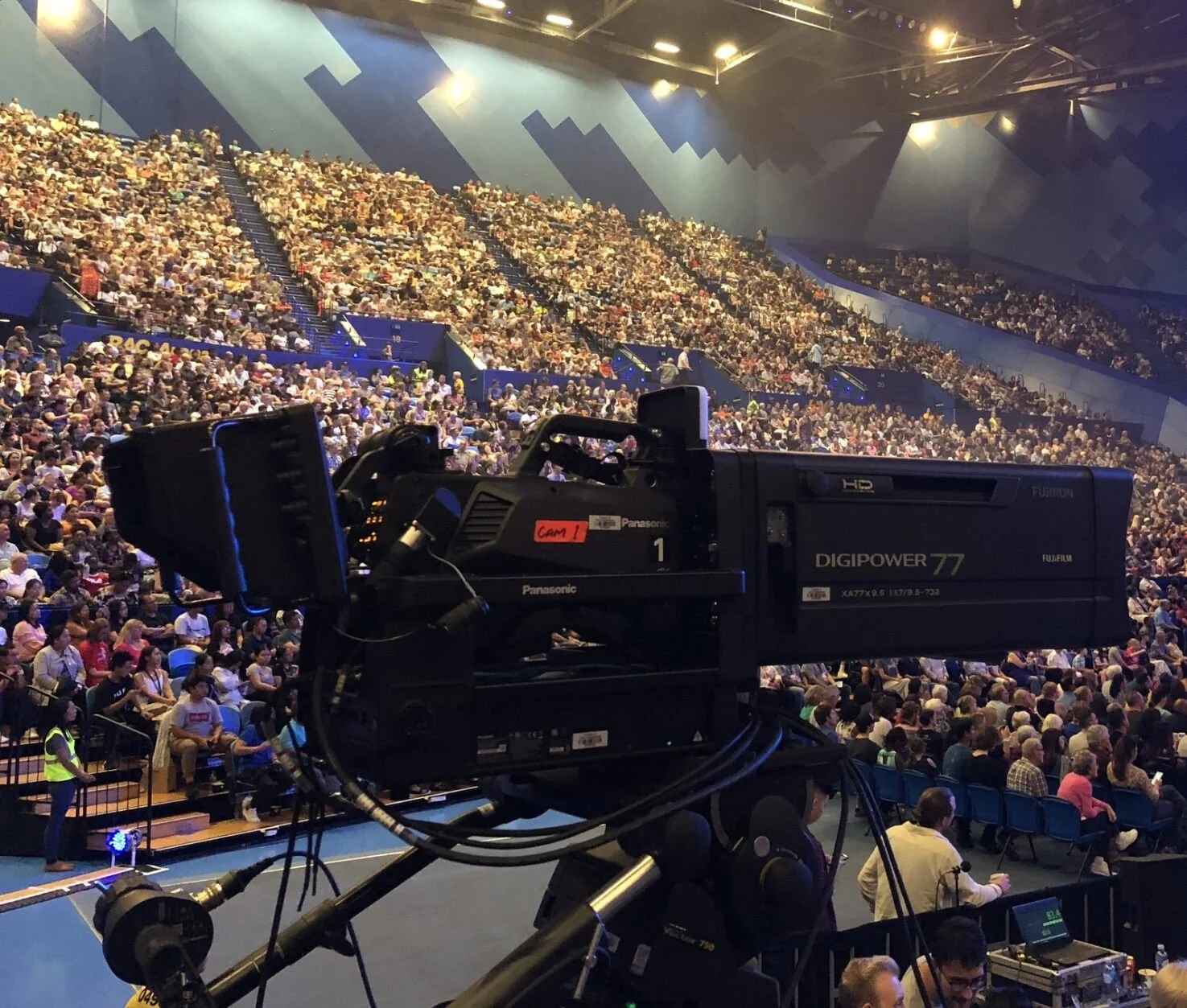 Professional video camera setup in a large indoor stadium filled with a seated audience.