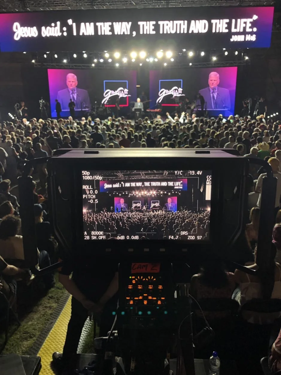 Large crowd attending an indoor religious event with a stage featuring a speaker, two large screens showing the speaker, and a prominent banner displaying a Bible verse."