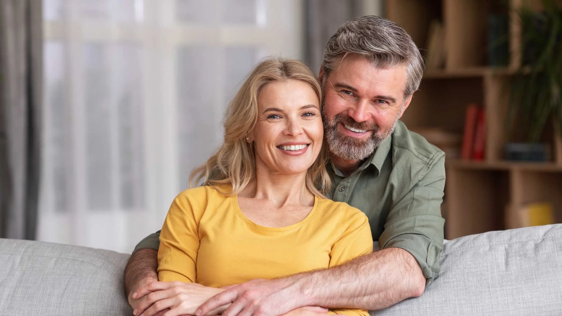 A happy middle-aged couple sitting on a couch, smiling at the camera, in a cozy living room.