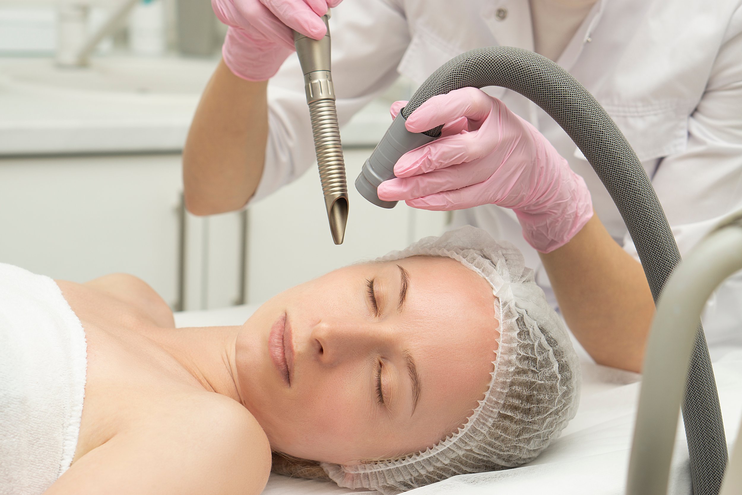 A woman receiving a facial treatment with a professional using a specialized device, both wearing gloves, in a clinical setting.