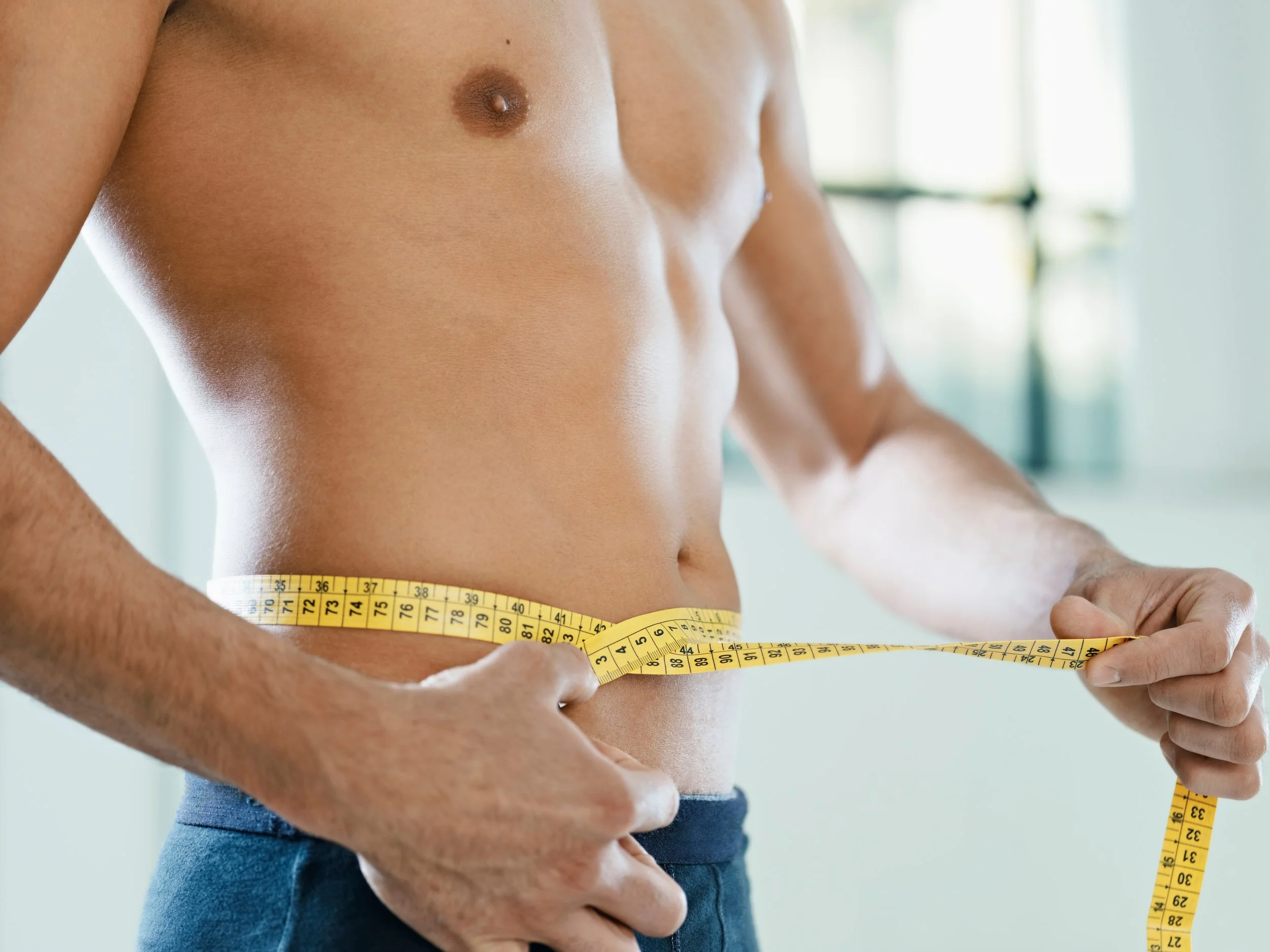 A shirtless man measuring his waist with a yellow tape measure in a bright room.