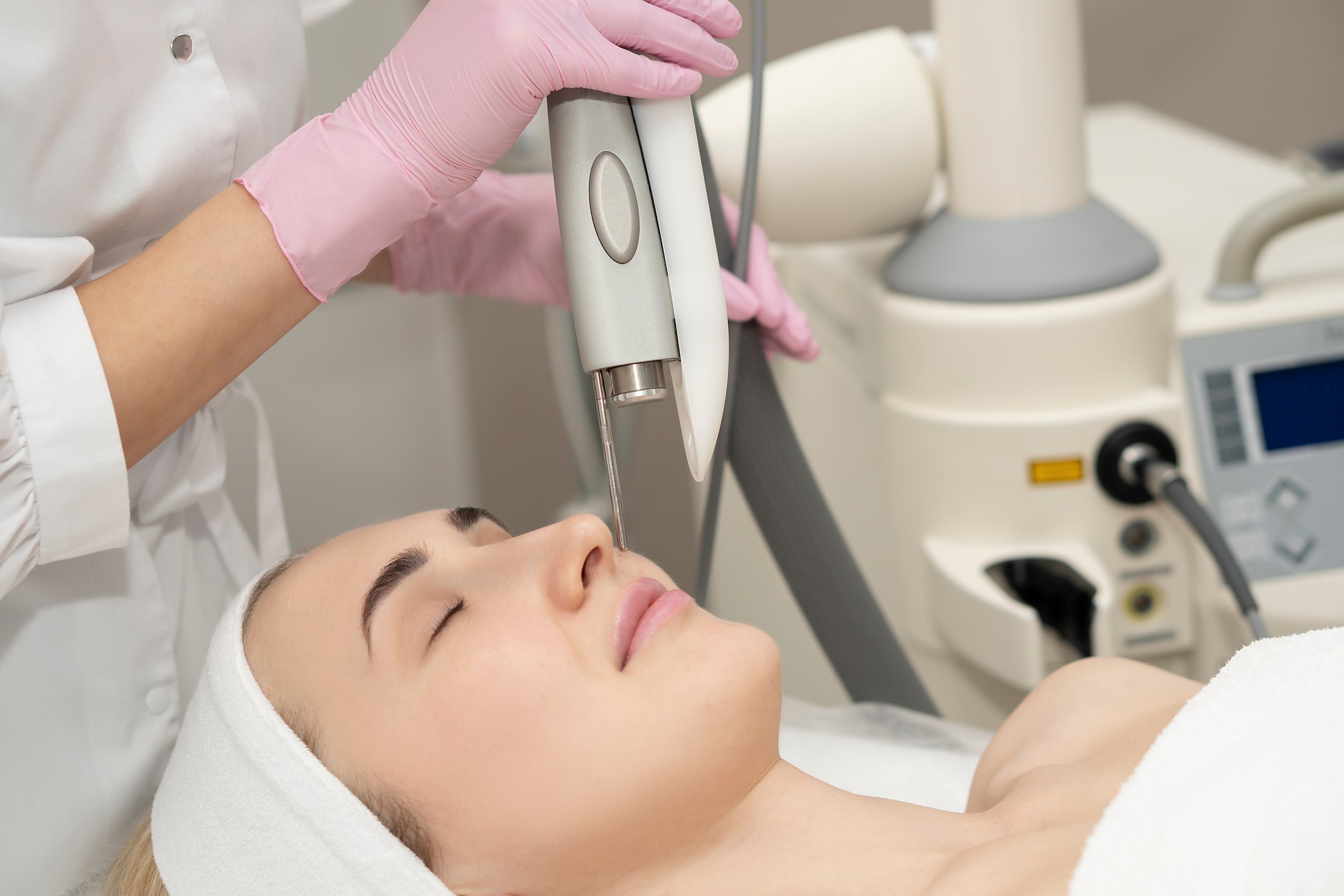 A woman receiving a facial treatment with a medical device in a clinic, lying on a bed with a towel wrapped around her head.