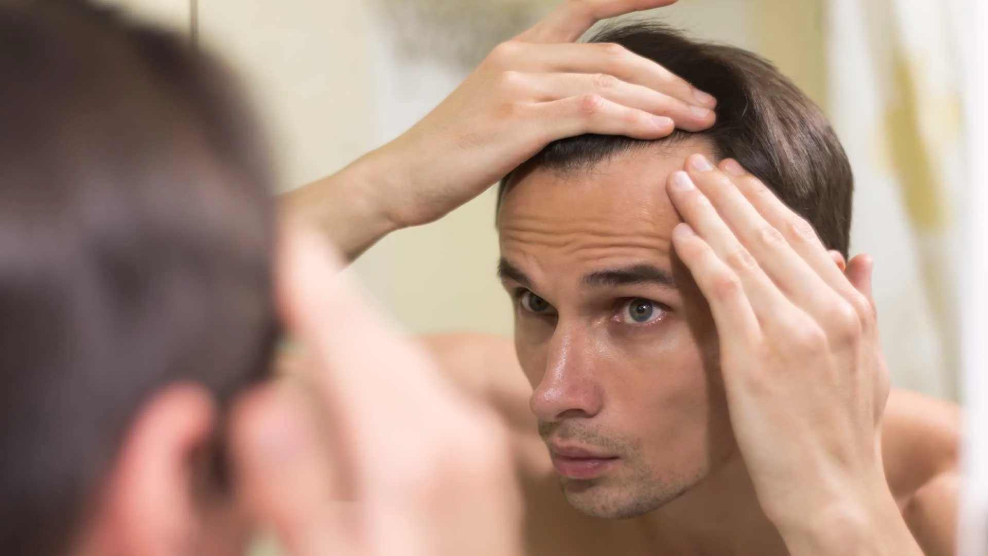 A man examines his hairline in a mirror, touching his scalp with both hands, noticing receding hair or thinning hair.