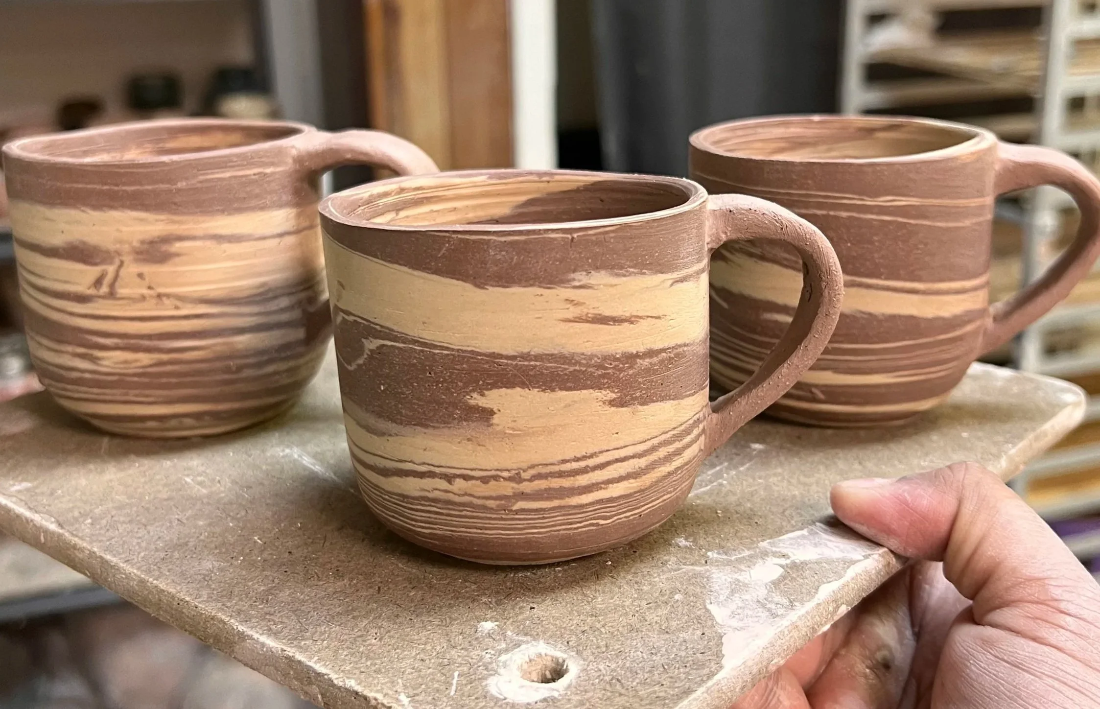Three handcrafted ceramic mugs with earthy tones and horizontal swirl patterns share a table in a pottery studio, with shelves of tools in the background.